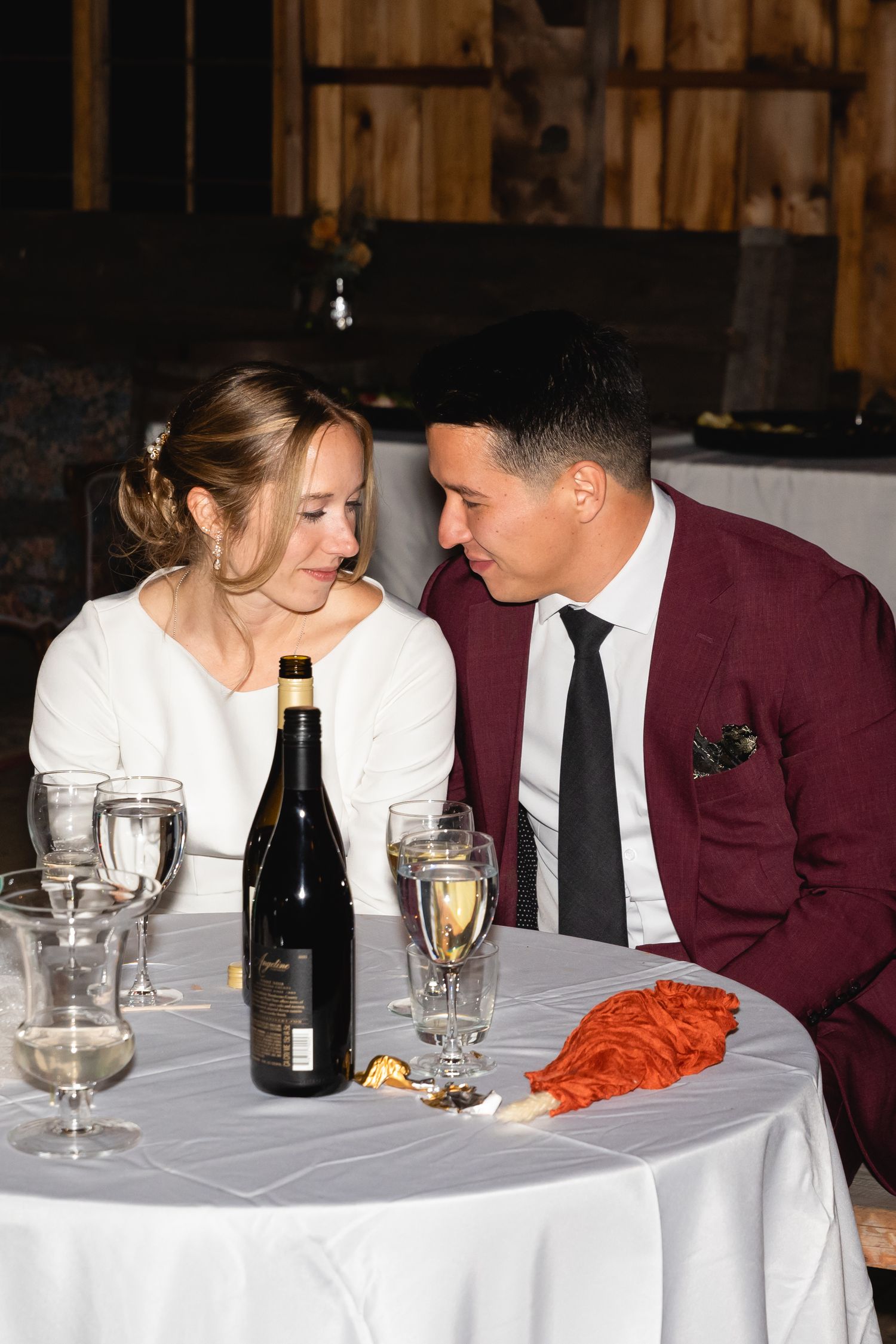 A couple shares an intimate moment over wine at a formal dinner table with white linens in a rustic venue.
