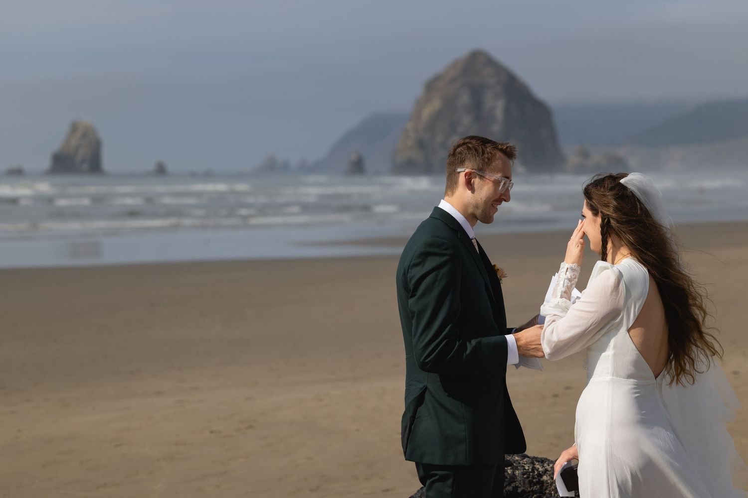 A couple shares an intimate moment on a misty beach with Haystack Rock visible in the background at Cannon Beach, Oregon.