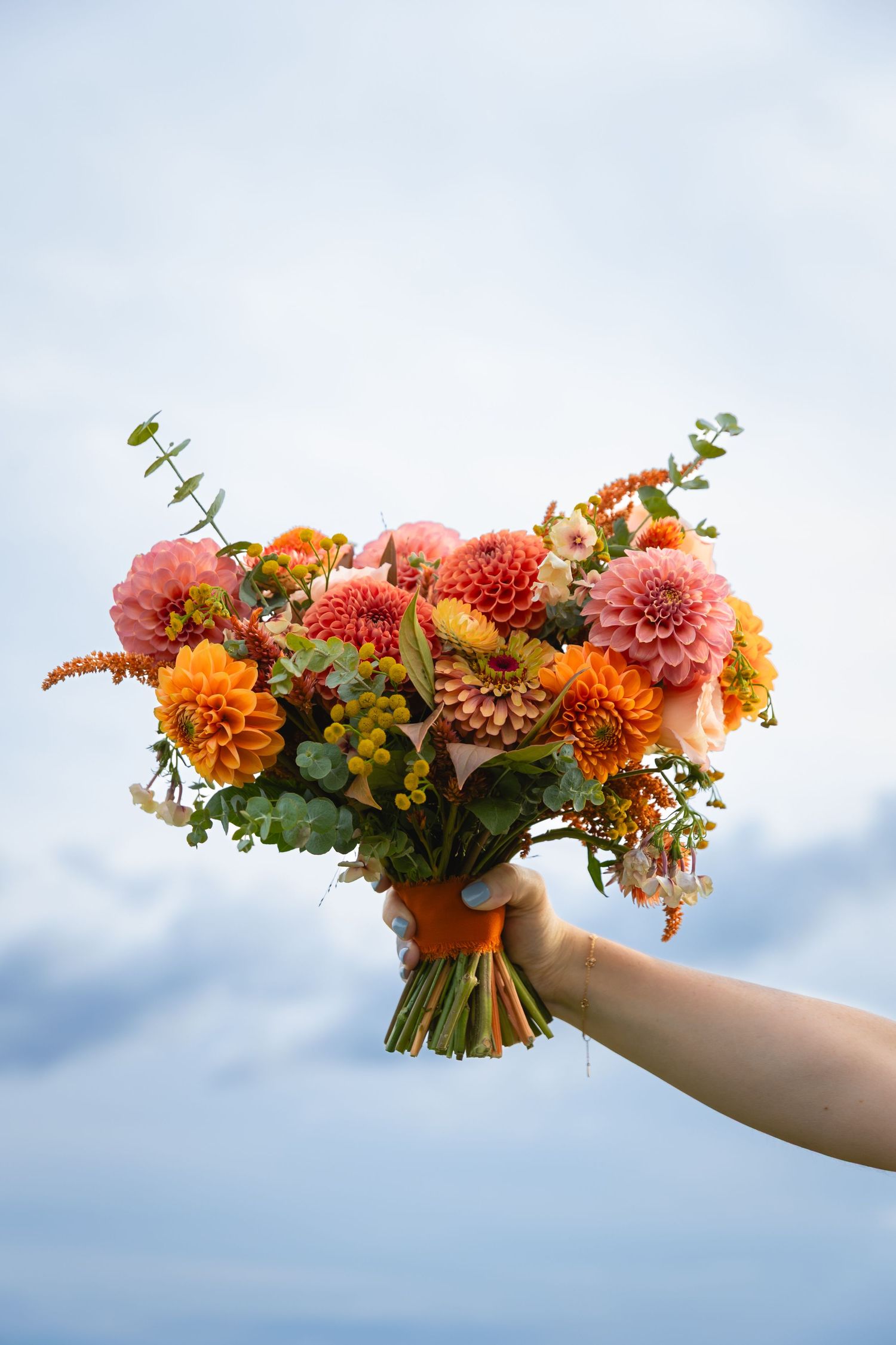 A vibrant coral and orange floral bouquet with dahlias and greenery extends horizontally across a blue sky background.