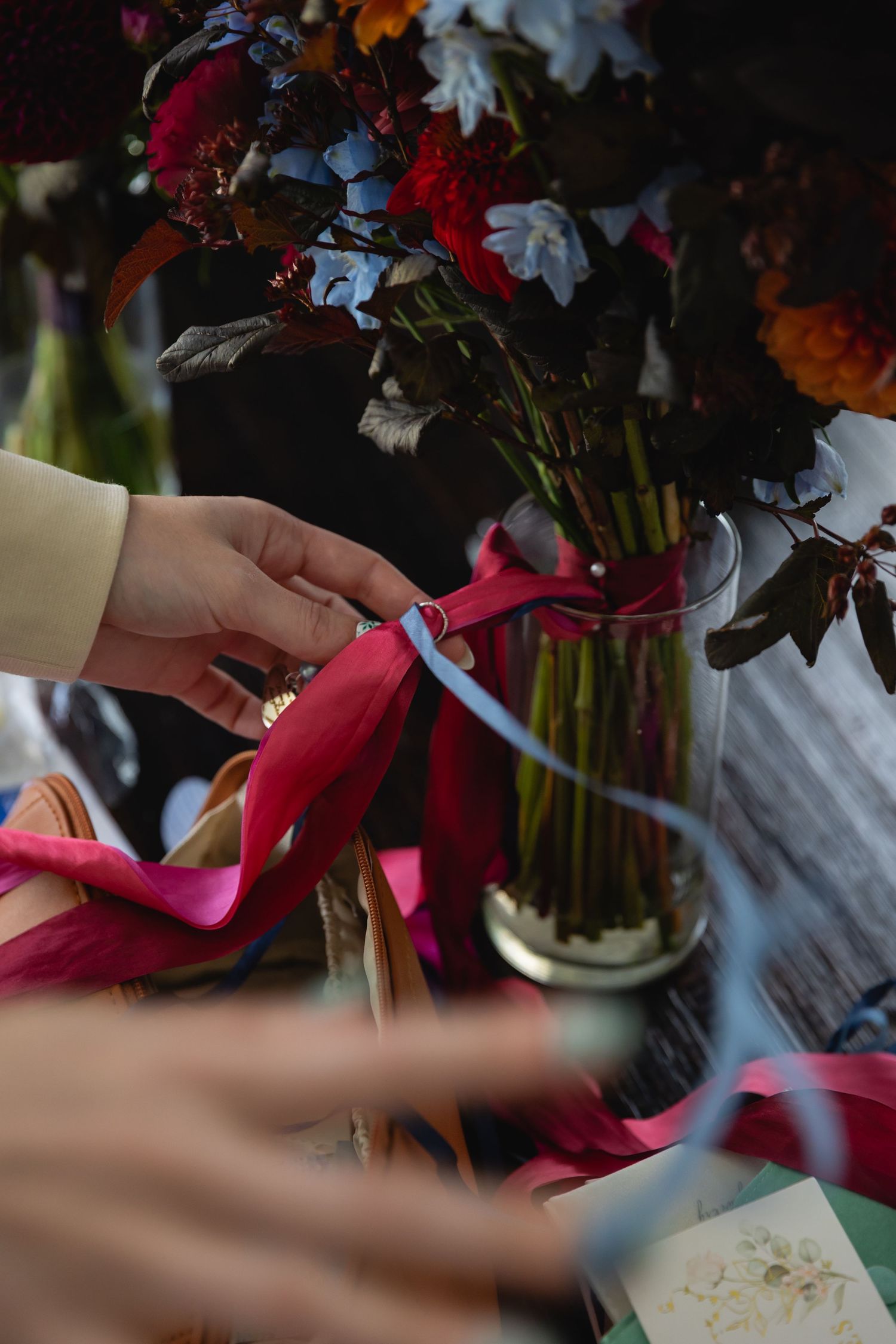 Hands arranging colorful flowers and ribbons in a vase with delicate care and precision.