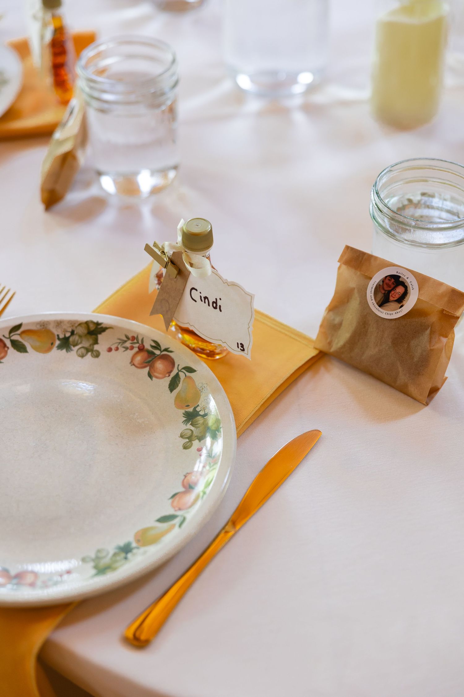 Place settings at a wedding reception with floral plates, brown paper bags, and orange napkins on white tablecloth.