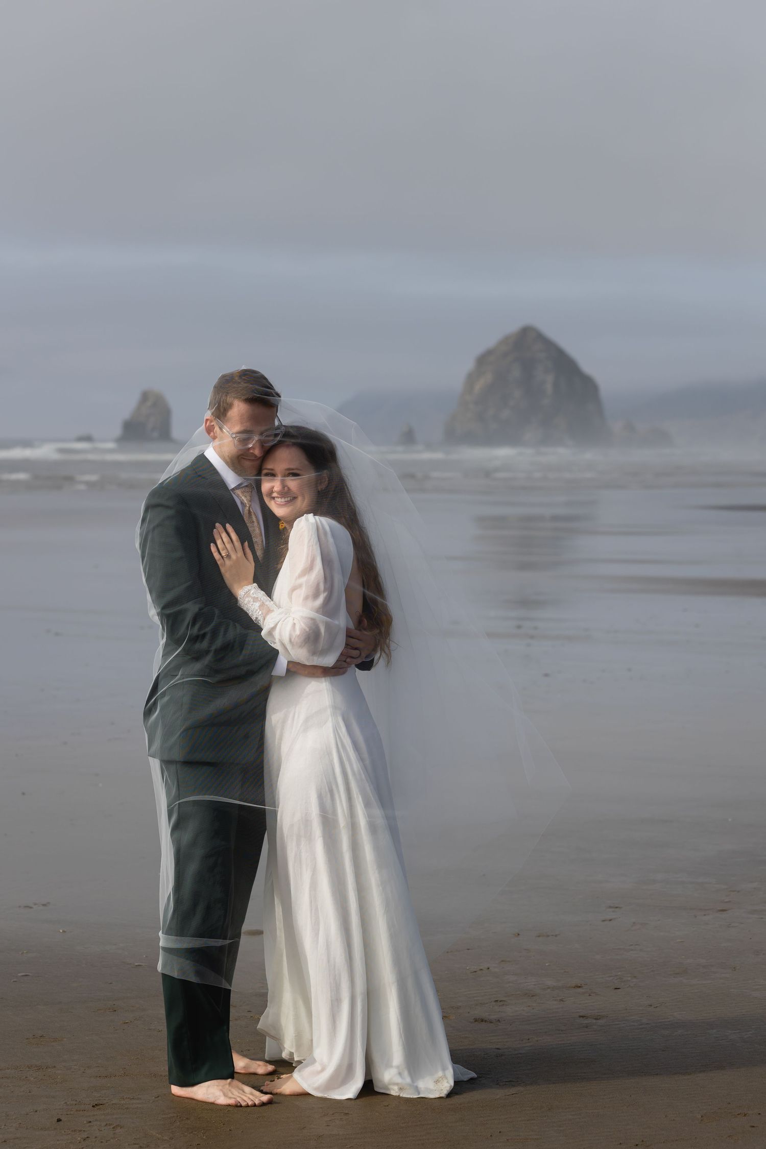 A couple in formal attire embraces on a misty beach with dramatic coastal rock formations in the background.