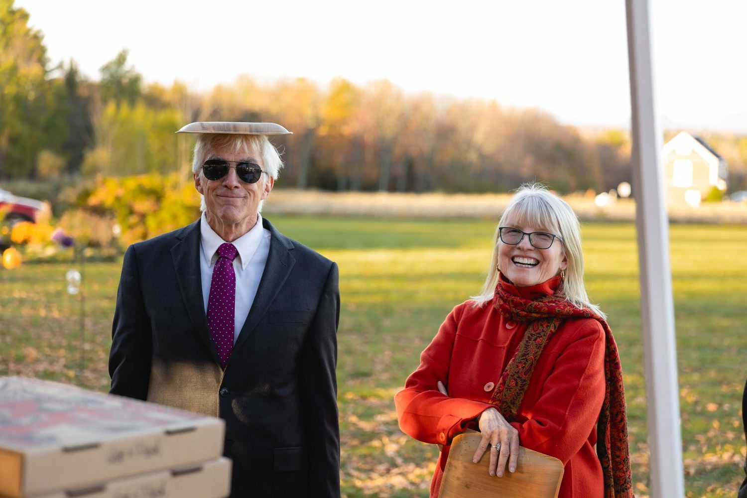 Two people stand outdoors in autumn, one wearing sunglasses and a suit, the other in a bright red jacket.