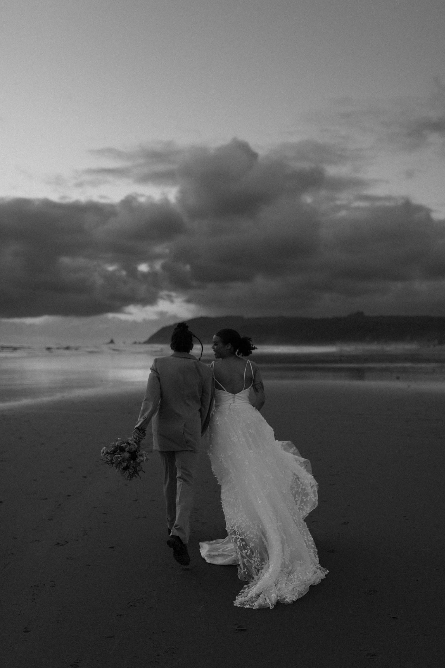 Black and white photo of newlyweds walking together on a moody beach at sunset.