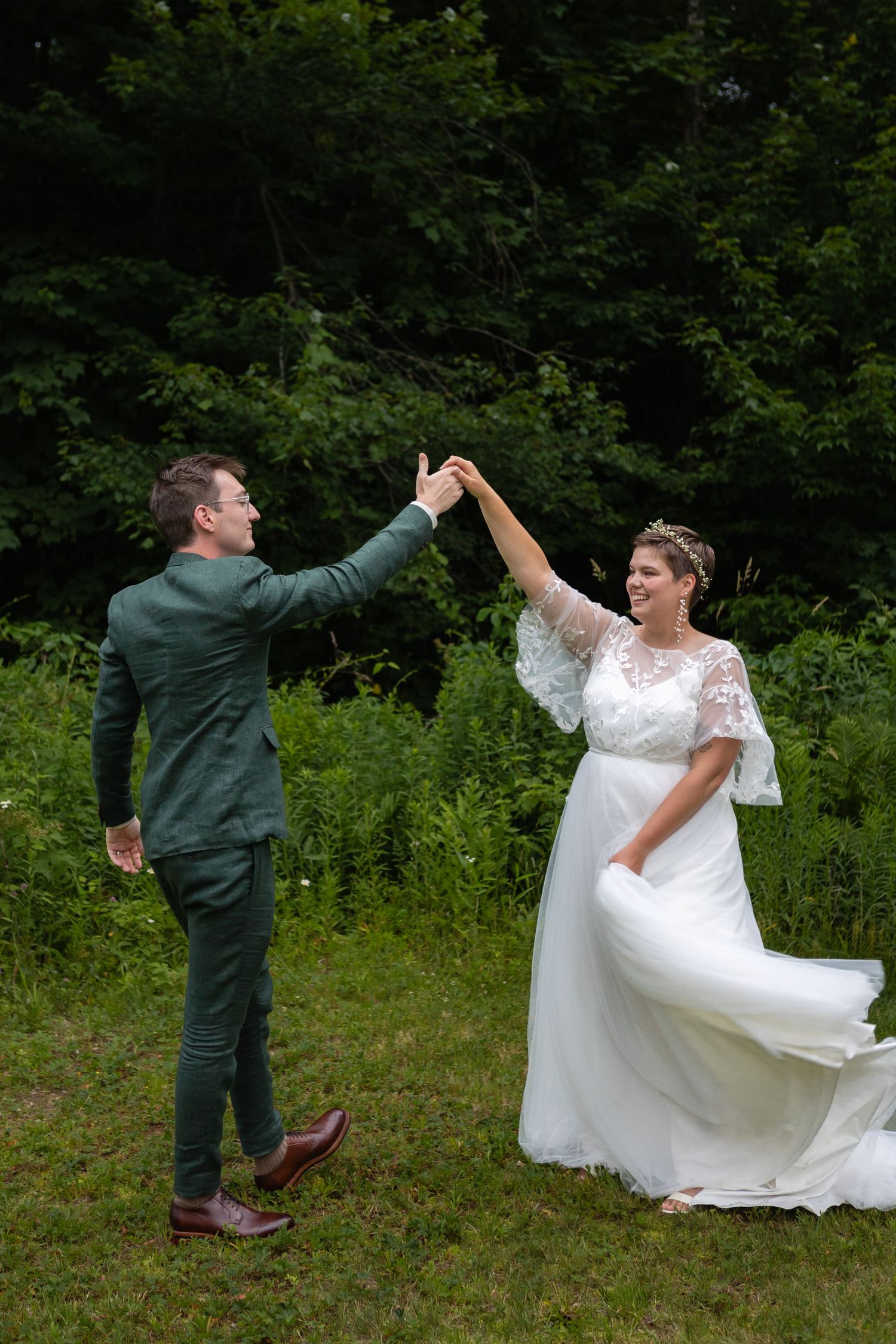 Bride in white dress and groom in green suit dancing playfully outdoors.