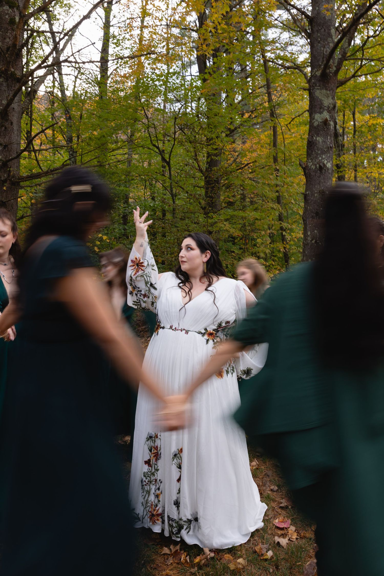 Bride in embroidered white dress celebrates with guests wearing dark green dresses in forest setting.