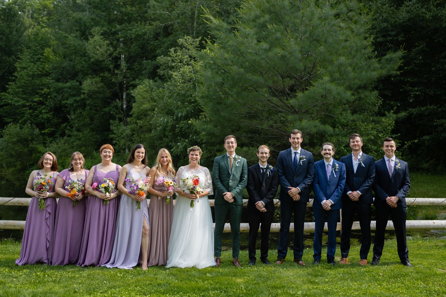 Wedding party in purple dresses and blue suits lined up for formal photo against forest backdrop.