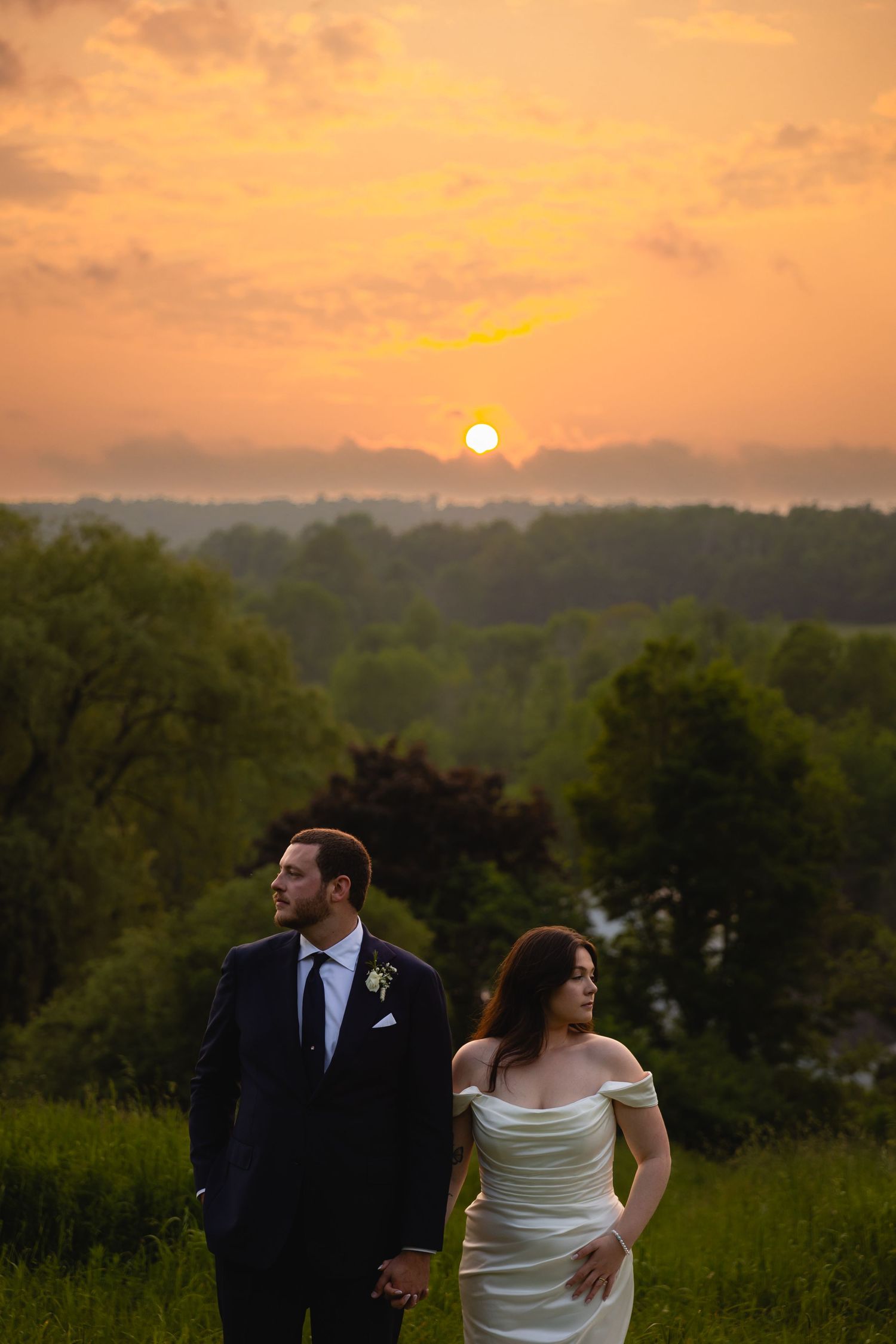 Bride and groom hold hands in lush green field beneath stunning golden sunset sky on wedding day.