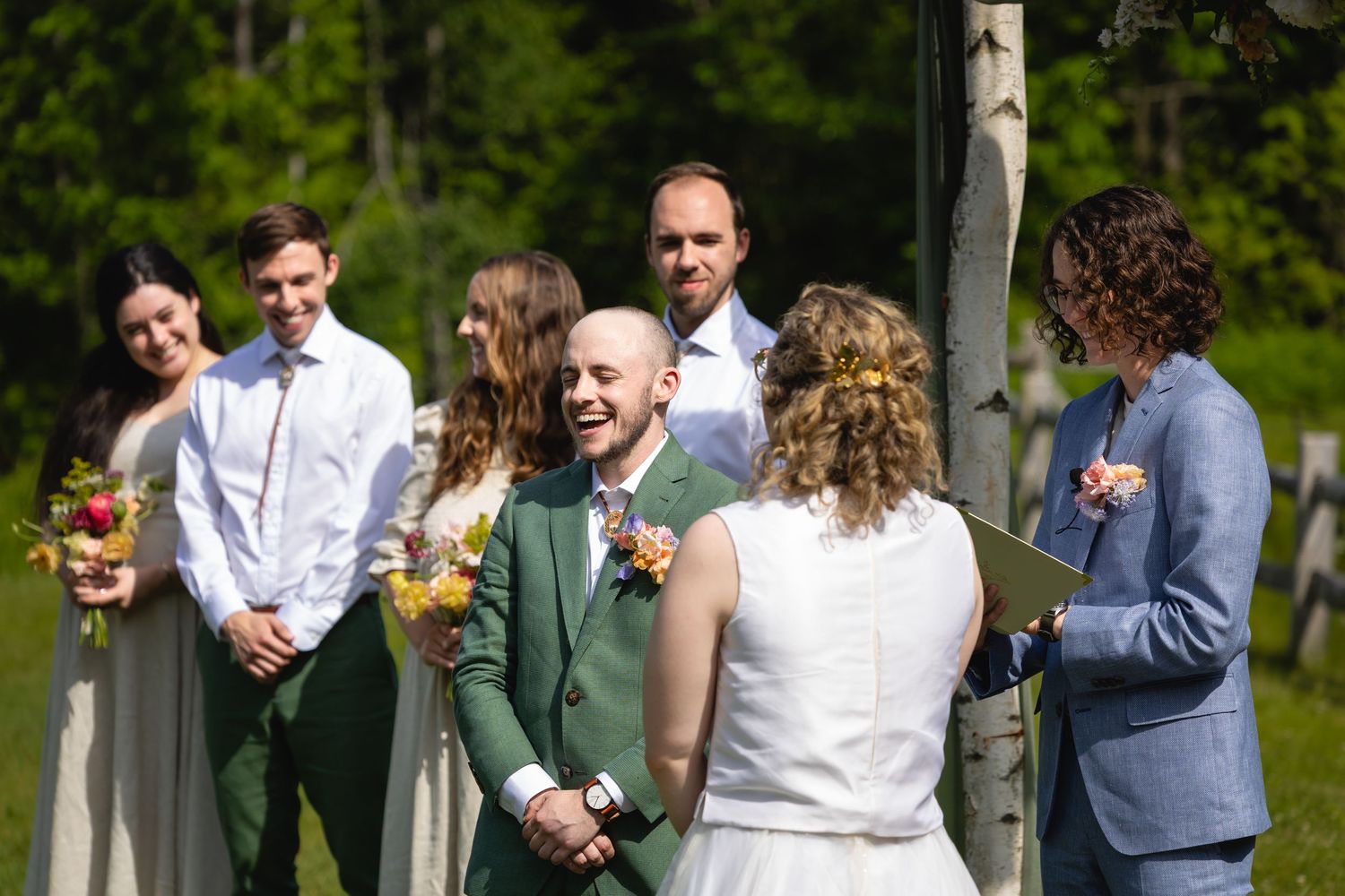 A small outdoor wedding ceremony takes place in a lush green setting with people gathered in casual attire.