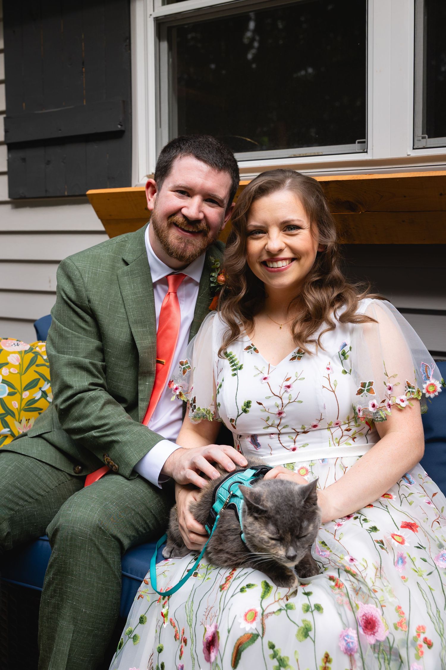 A couple in formal attire sits outdoors with a gray cat wearing a teal collar.