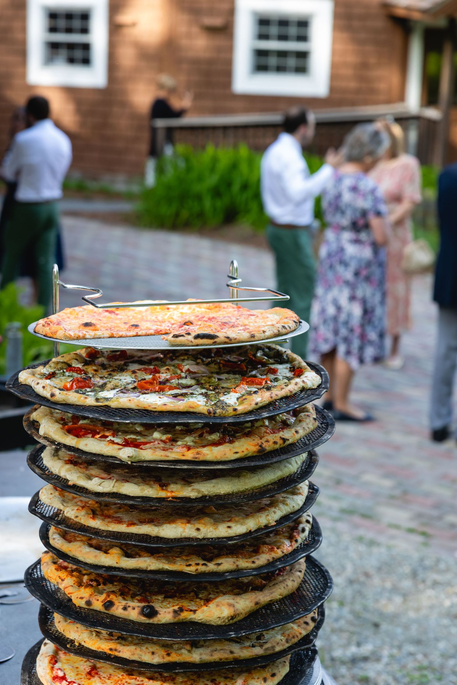 A tall stack of colorful pizzas on metal serving trays at an outdoor event with guests in the background.
