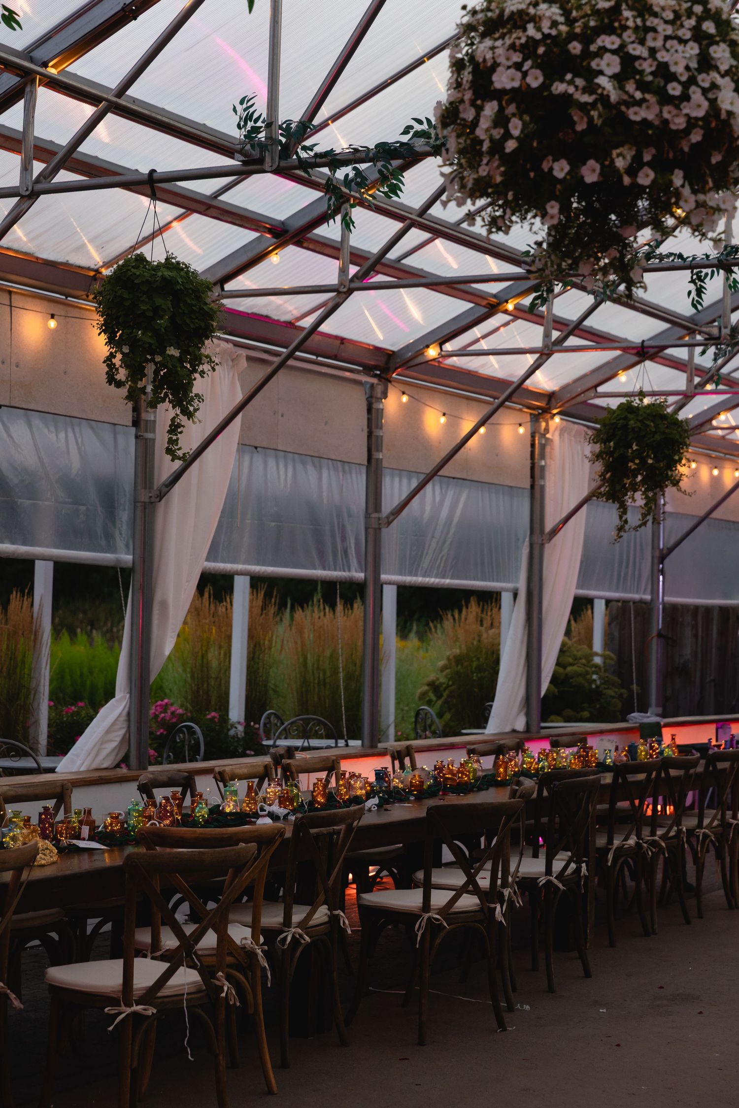 Long rustic wooden dining tables set up inside a clear tent structure with string lights and floral arrangements at sunset.