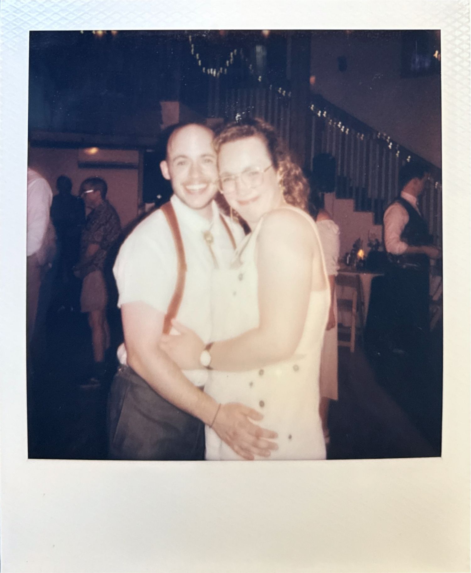 A vintage Polaroid photo from a social gathering shows a couple embracing and smiling at the camera in an indoor setting.