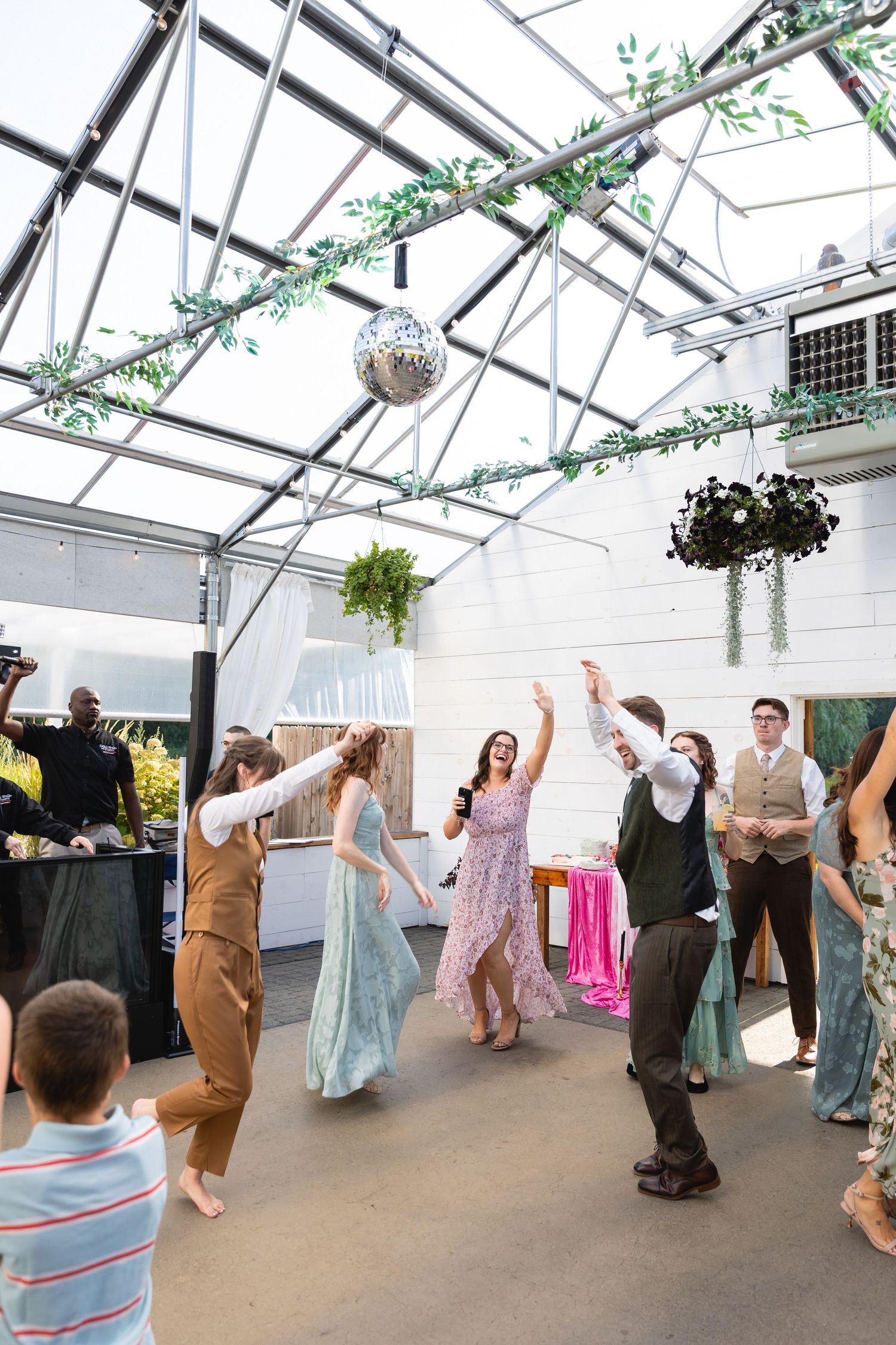 Wedding guests dance in a line formation under a white greenhouse-style tent during a celebration.