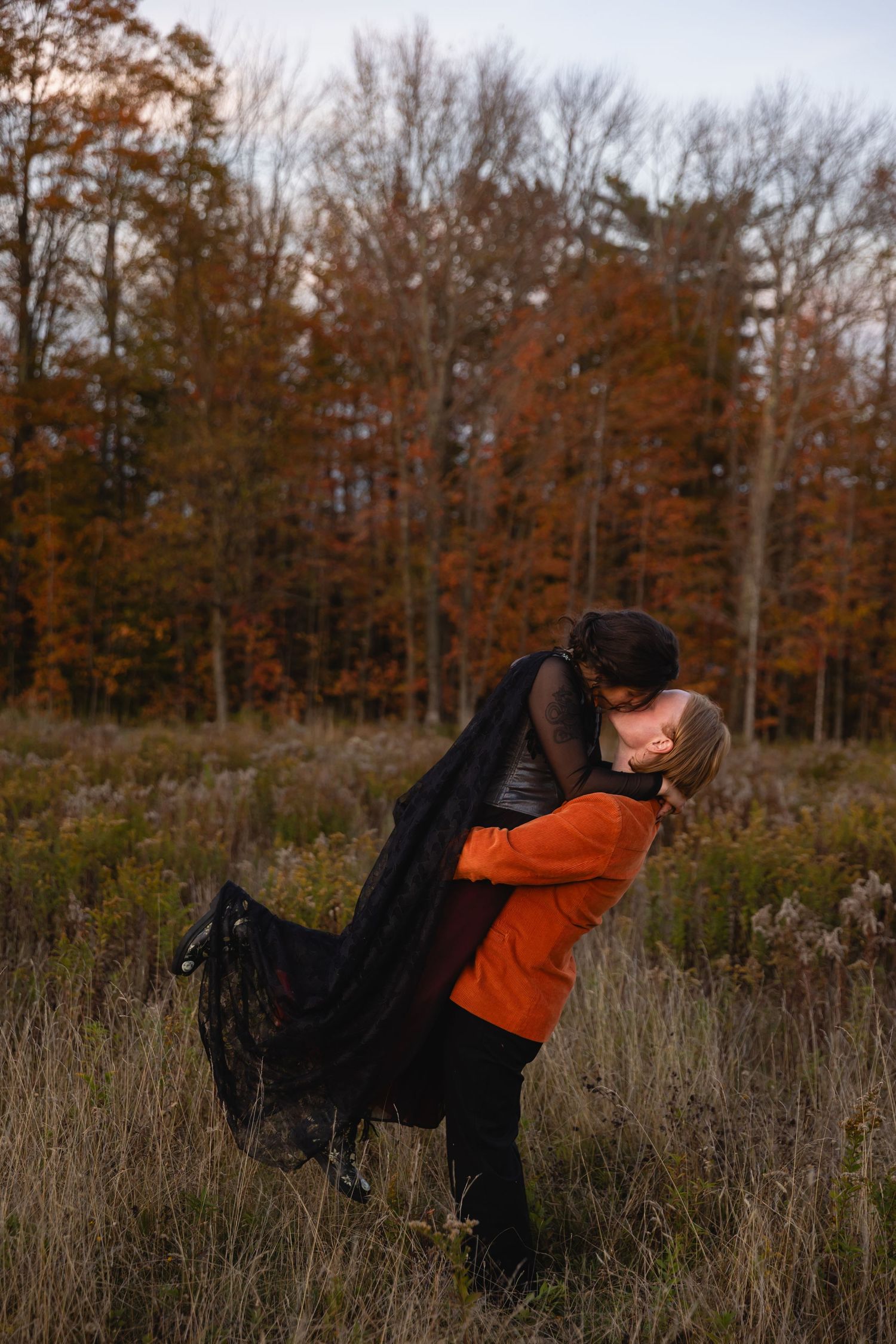 A couple embraces in a romantic dance pose against an autumn forest backdrop with tall grass and warm orange foliage.