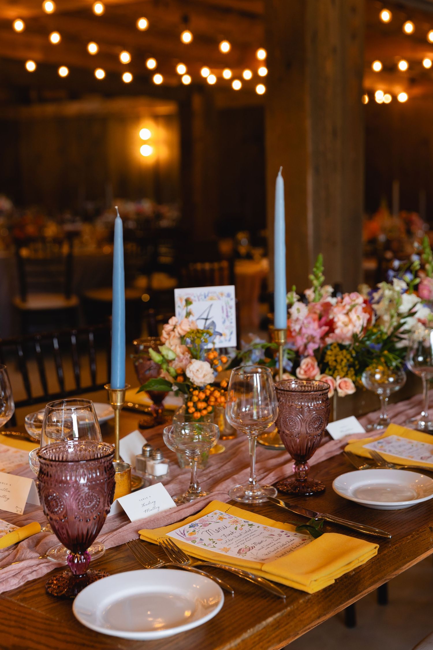 Elegant table setting with blue candles, pink flowers and vintage purple glassware at rustic wedding reception.