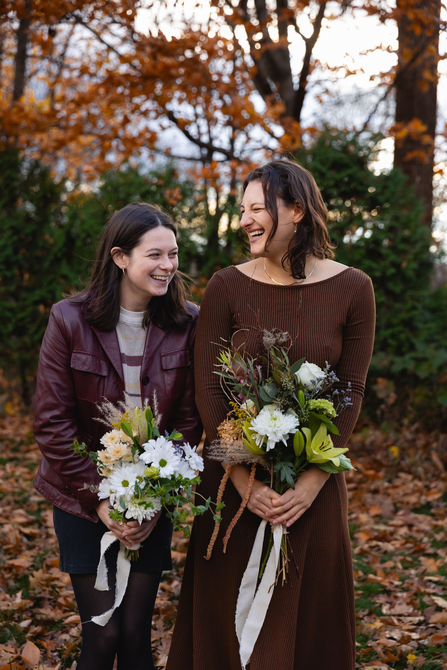Two people share laughter while holding beautiful floral bouquets on an autumn day with fallen leaves and trees.