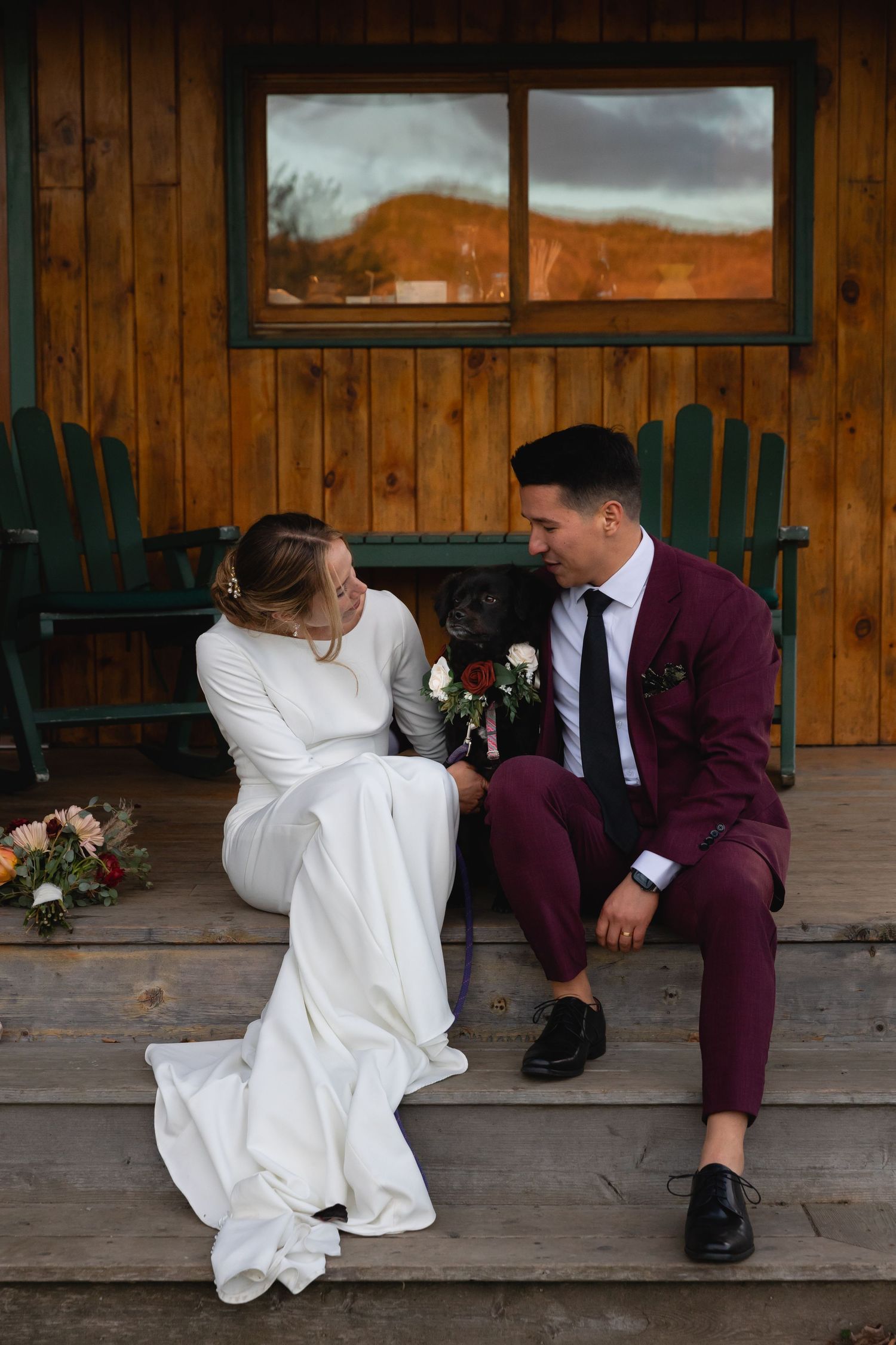 A couple sits on a rustic wooden porch sharing an intimate moment, the bride in white and groom in burgundy suit.