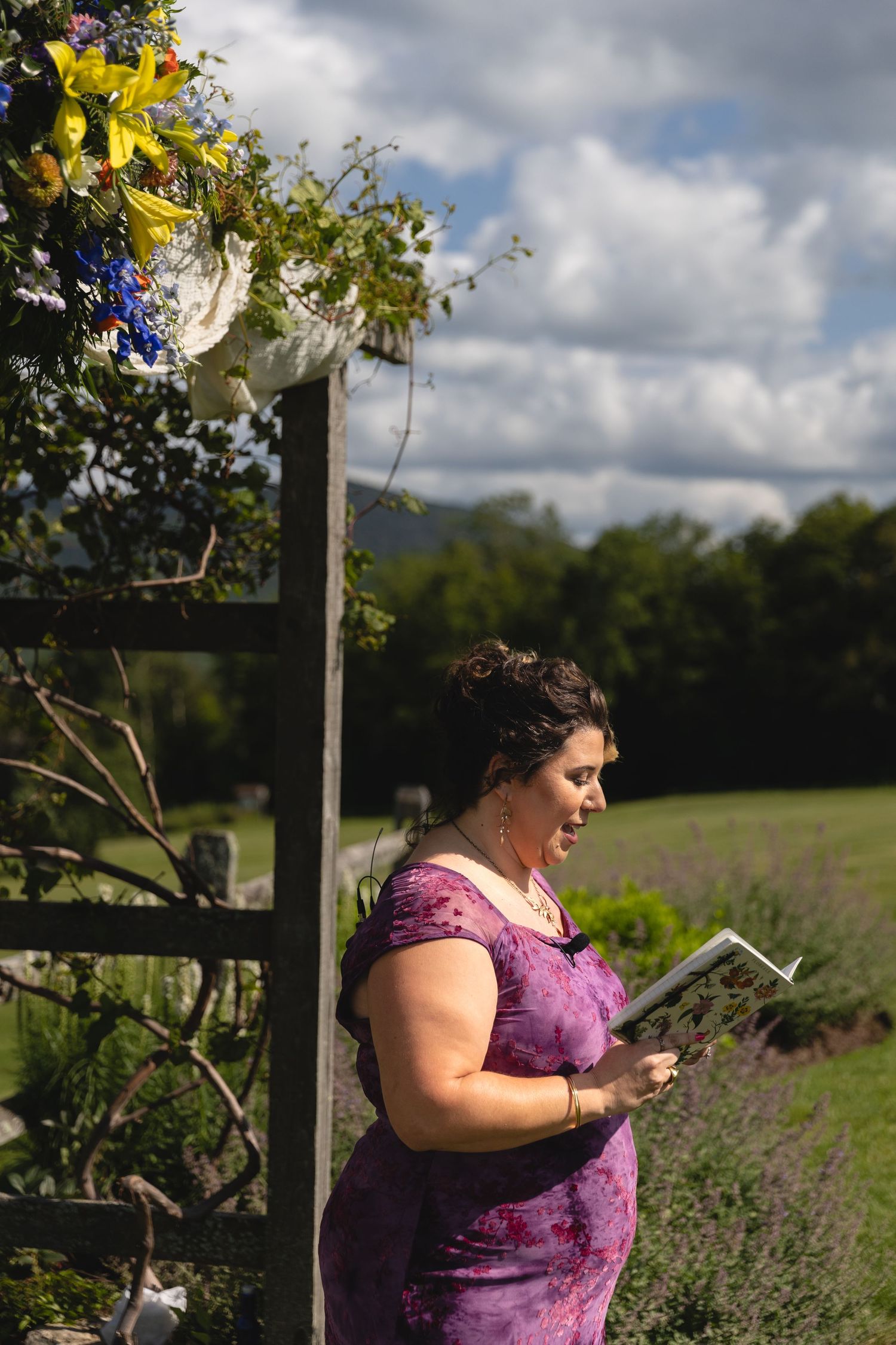 A person in purple reads from a document under a floral archway at an outdoor venue.