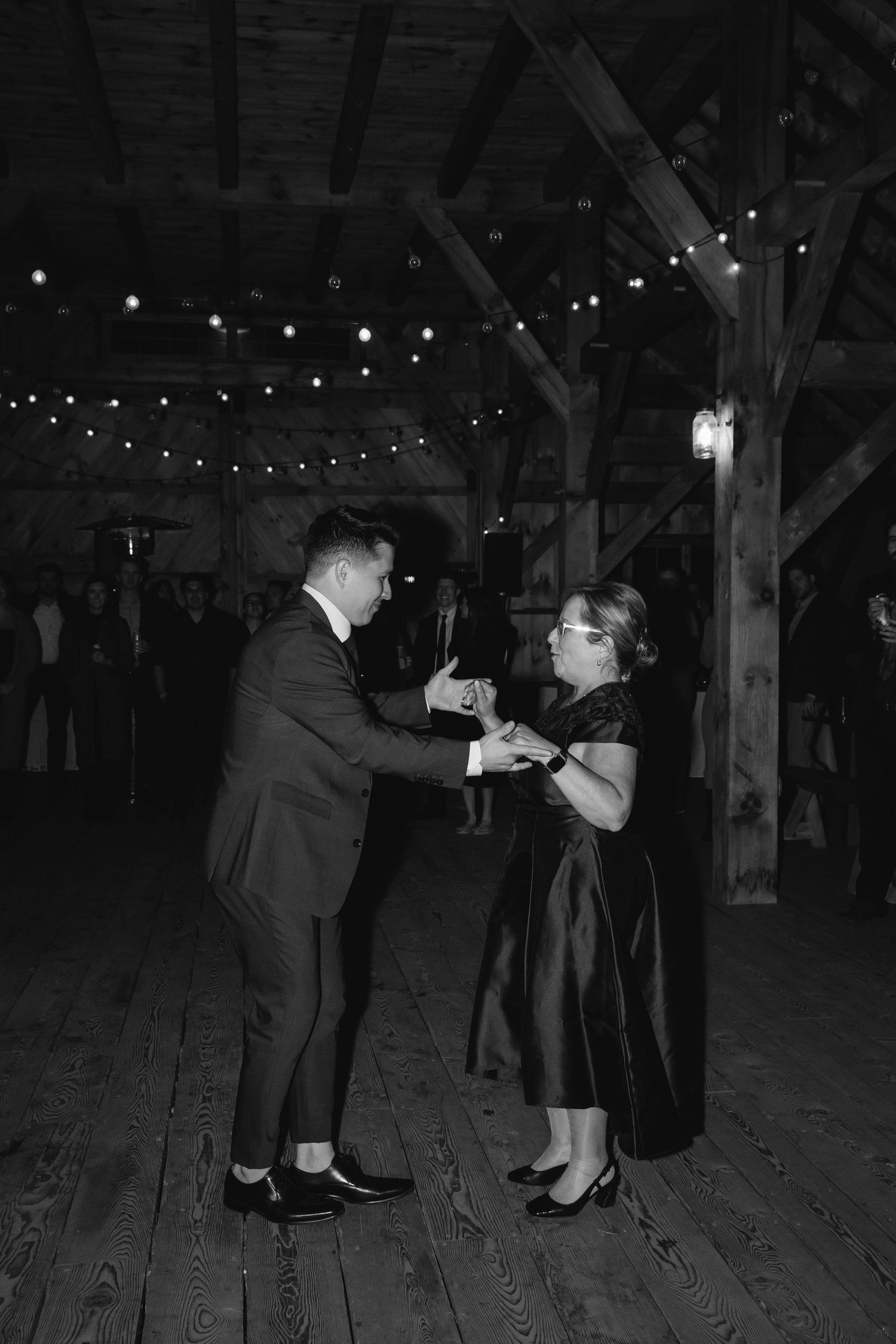 A couple dances together in a rustic barn venue lit by string lights and wooden beams.