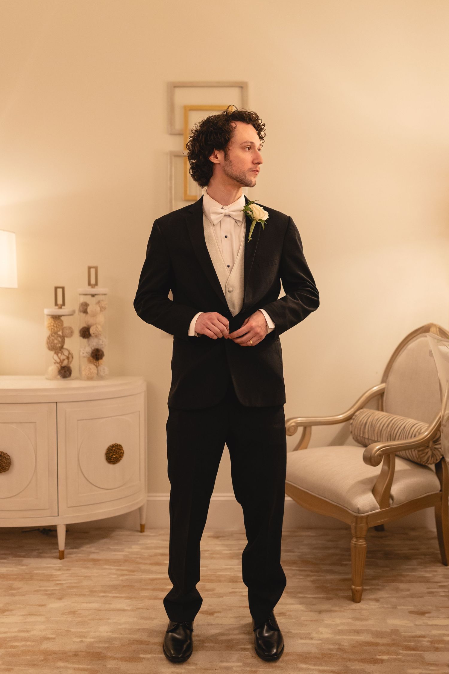 Groom in classic black tuxedo with white bow tie and boutonniere poses in elegant room with vintage furniture.