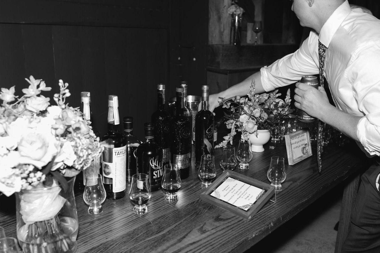 A bartender works behind a wooden bar counter with bottles and floral arrangements in black and white.
