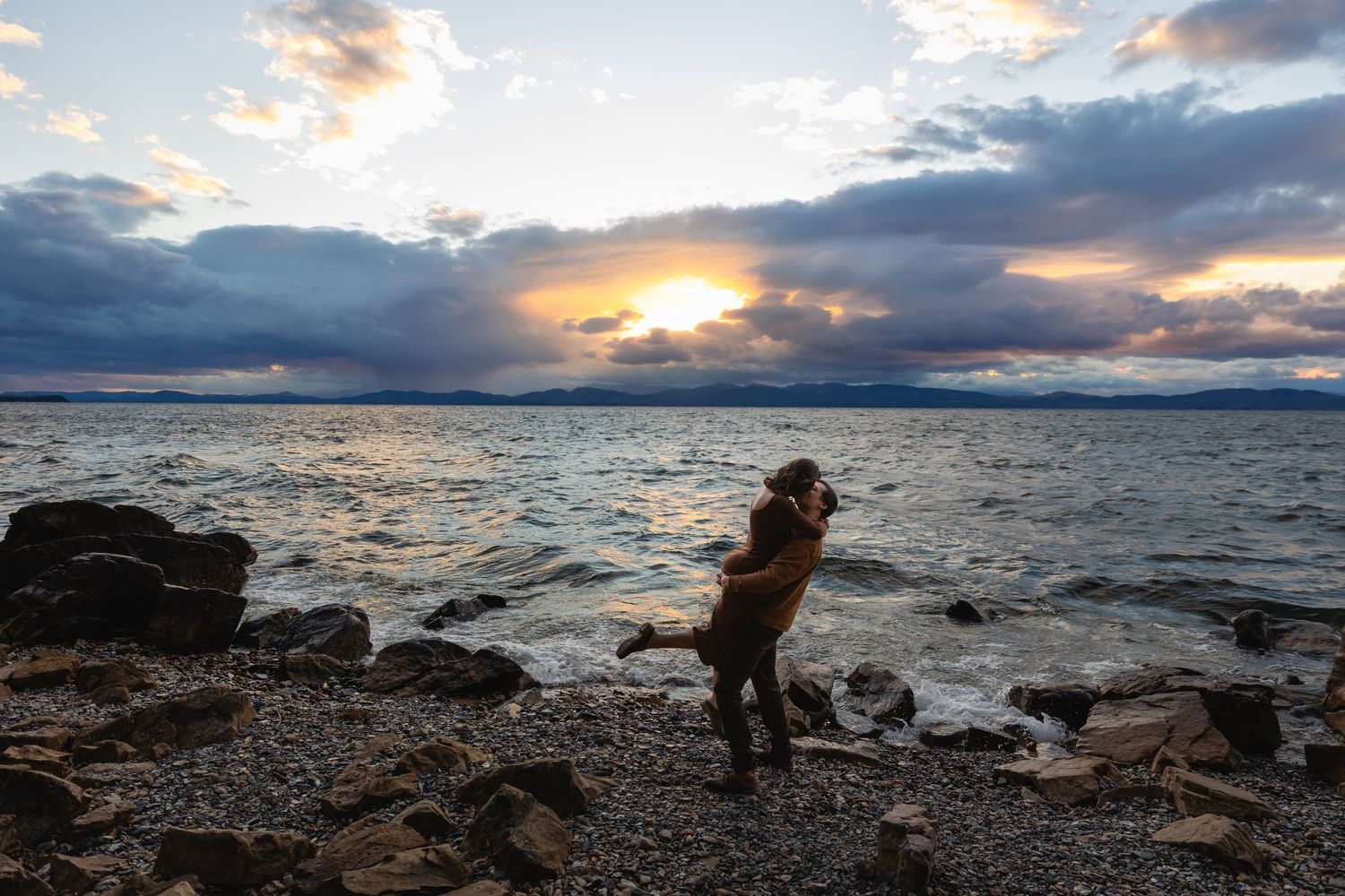 A dog stands on a rocky beach at sunset with dramatic clouds and mountains in the background as waves roll in.