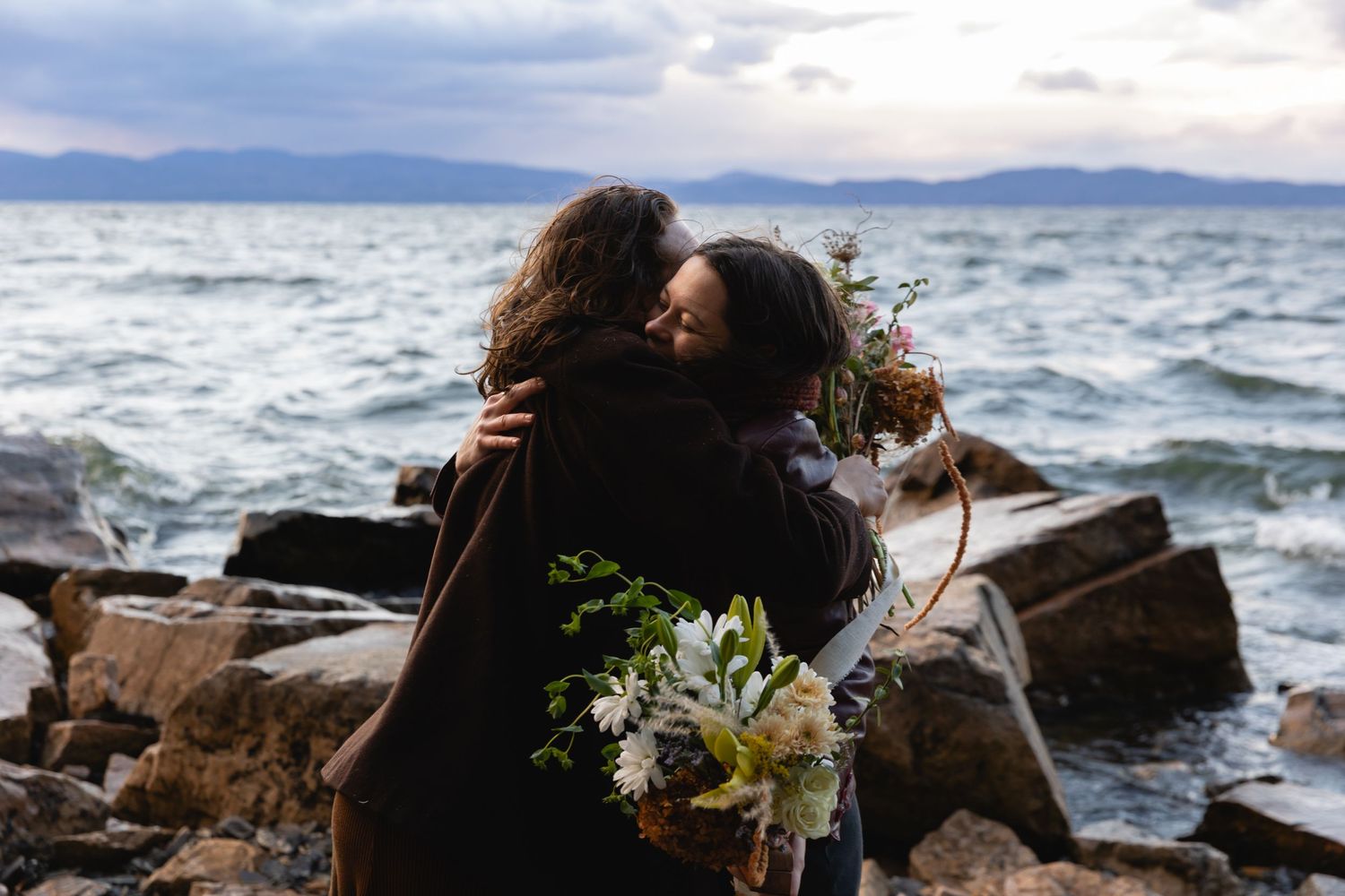 Two people embrace on a rocky coastline with a bouquet of white flowers and stormy ocean waters in the background.