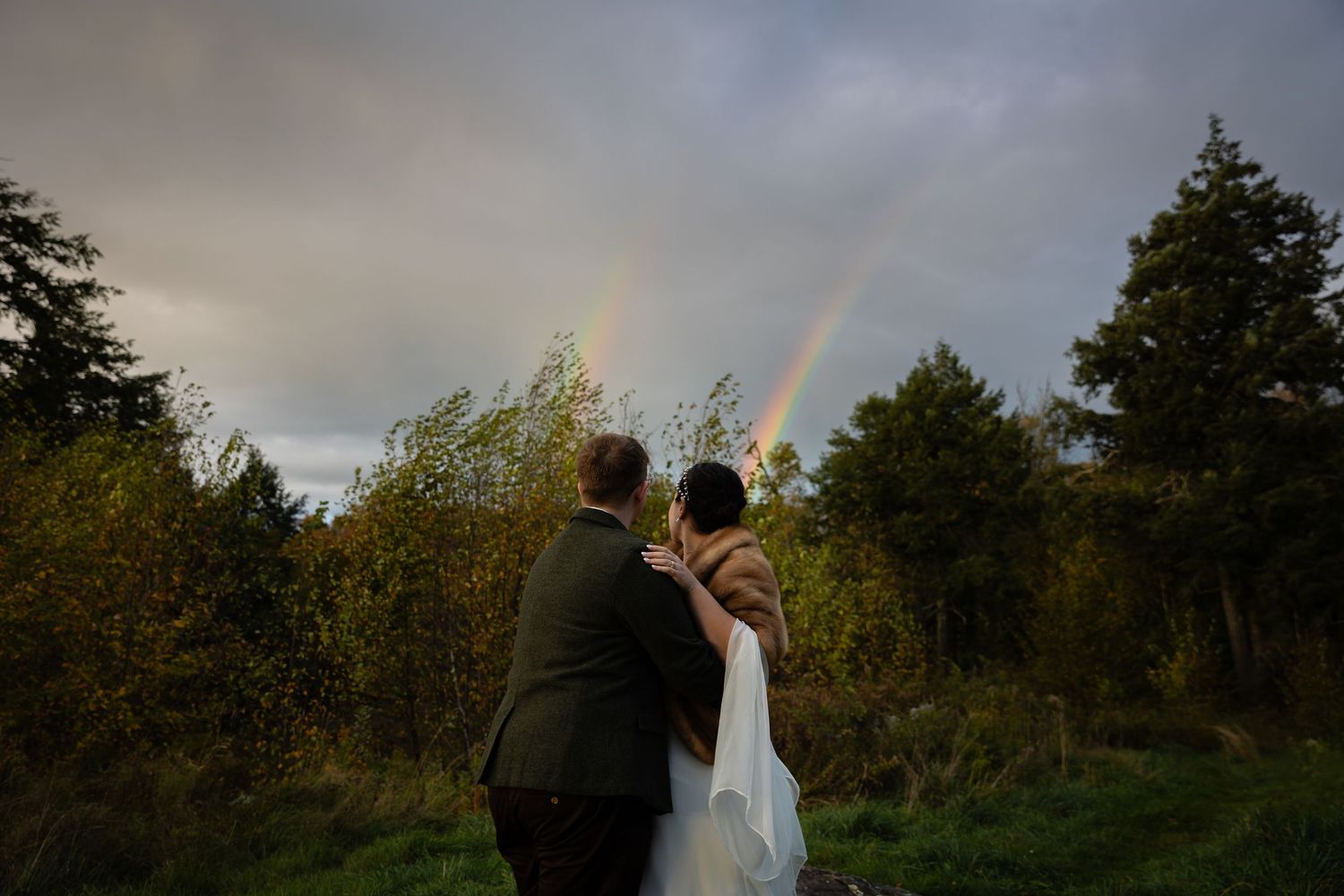 Newlywed couple admires double rainbow over lush green landscape after outdoor wedding.