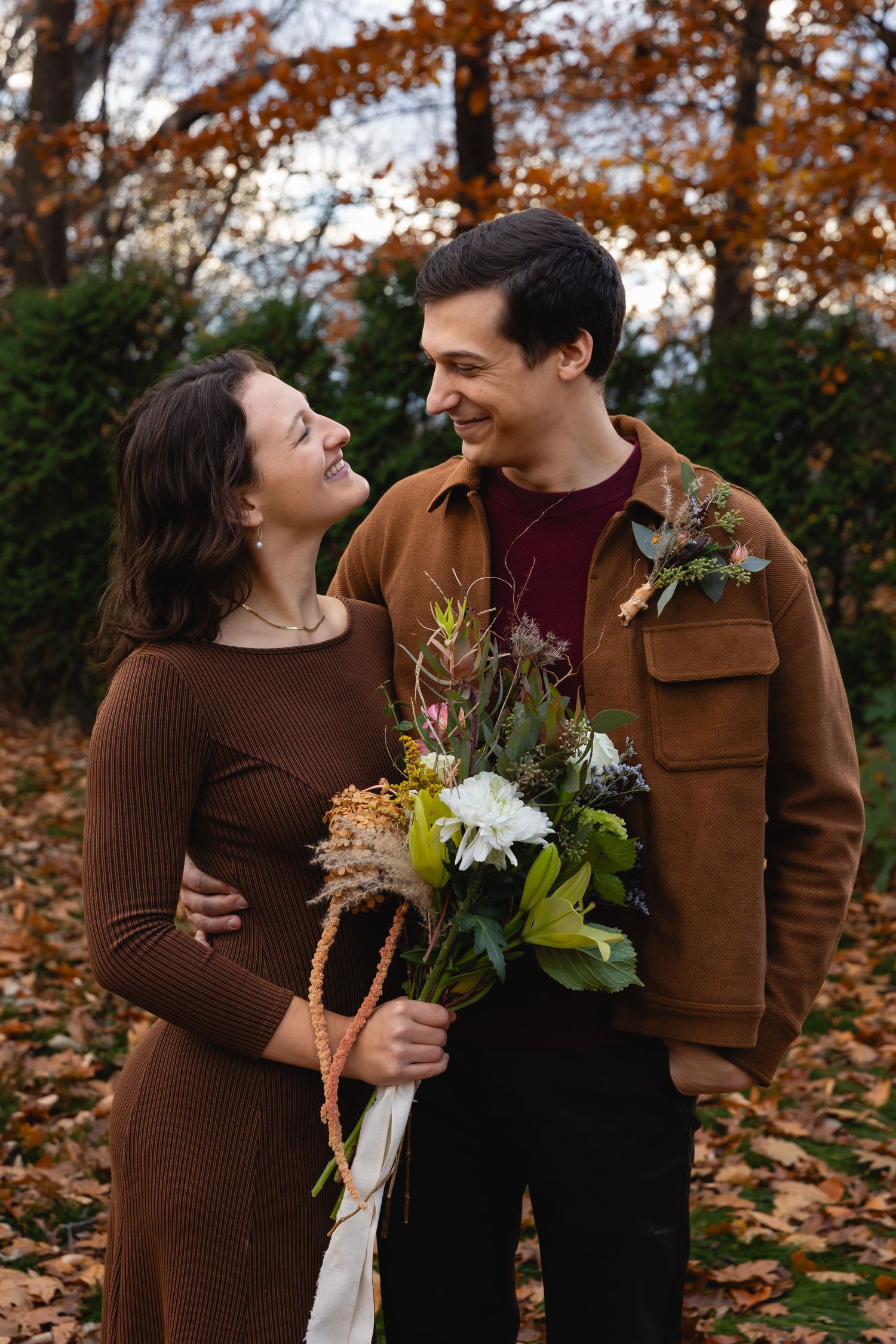 A couple shares a romantic moment outdoors among fall foliage while holding a bouquet of fresh flowers.
