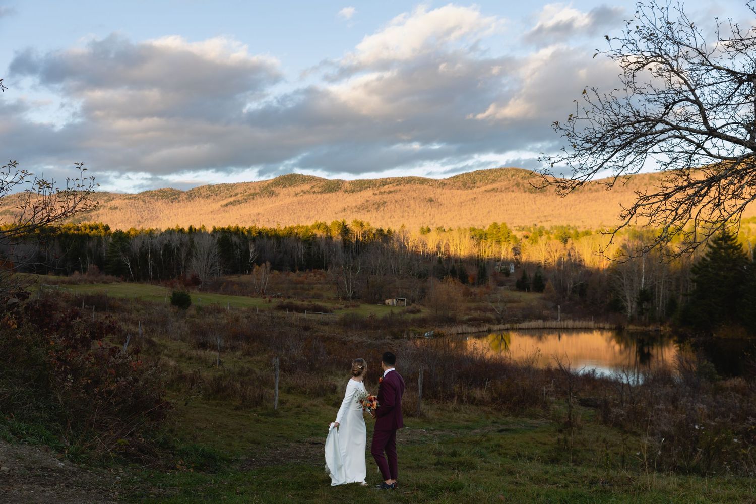 Two figures in formal attire stand together against a scenic autumn mountain landscape with a serene lake at sunset.