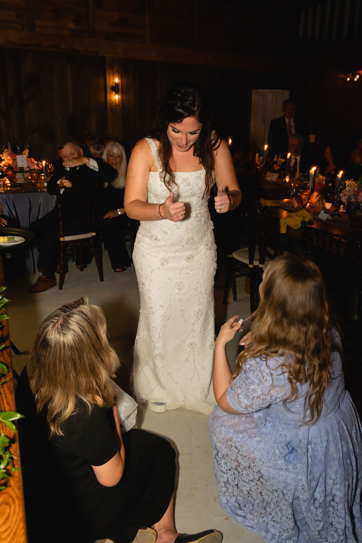 A bride in a fitted white lace gown dances with guests at a dimly lit wedding reception with candles and table settings.