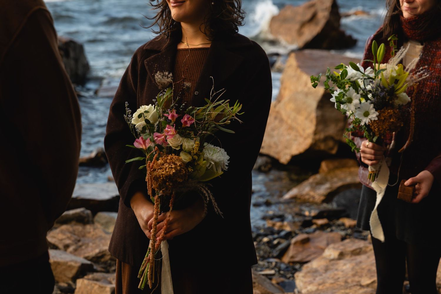 People in black attire holding colorful floral bouquets stand on a rocky shoreline at sunset.
