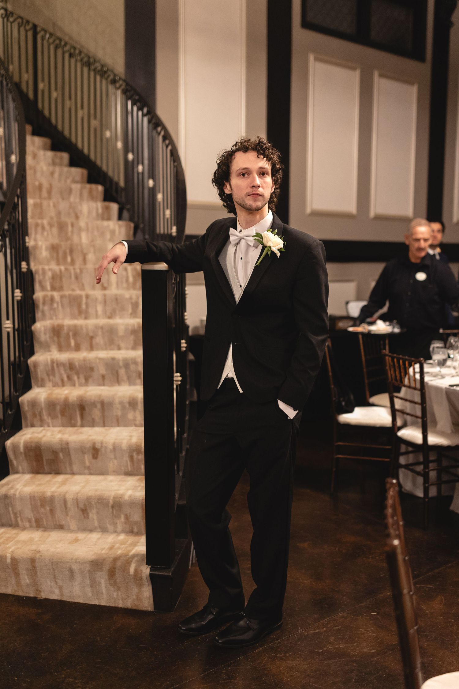 Classic groom in black tuxedo and white boutonniere stands casually by elegant staircase at wedding reception.