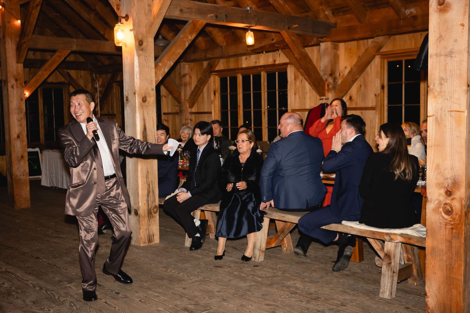 People gathered in a rustic wooden barn venue with wooden beams and benches for an indoor celebration.