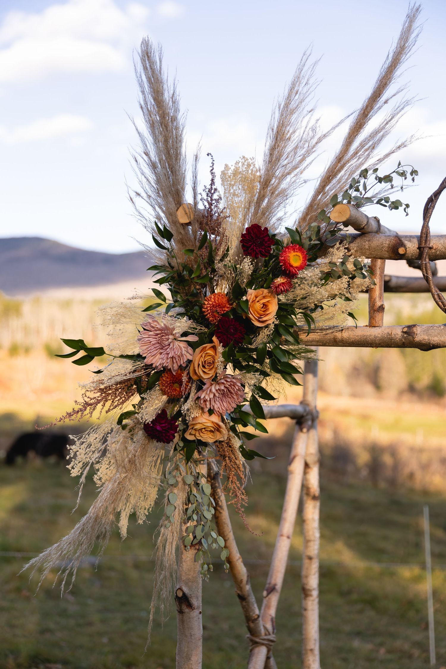 A rustic autumn floral arrangement with pampas grass and roses decorates a birch arch in a pastoral mountain landscape.