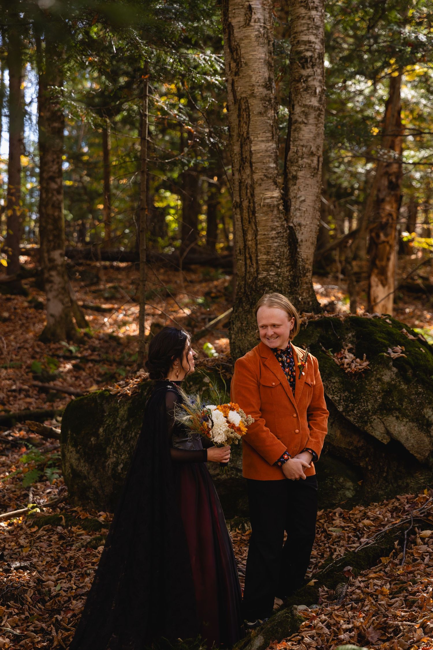 Two people stand close together in a wooded area during autumn, surrounded by fallen leaves and tall trees.