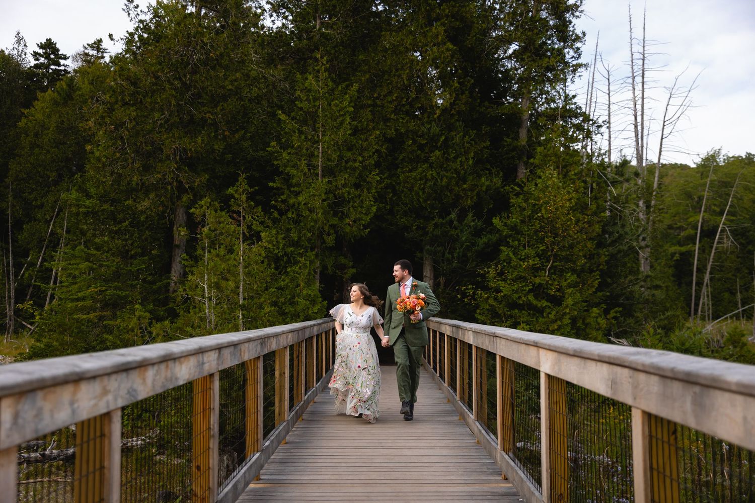 A couple in wedding attire walks together on a wooden bridge surrounded by lush forest trees.