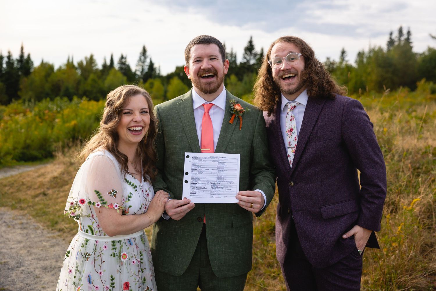 A group laughs together outdoors during a wedding celebration in a scenic natural setting with greenery.