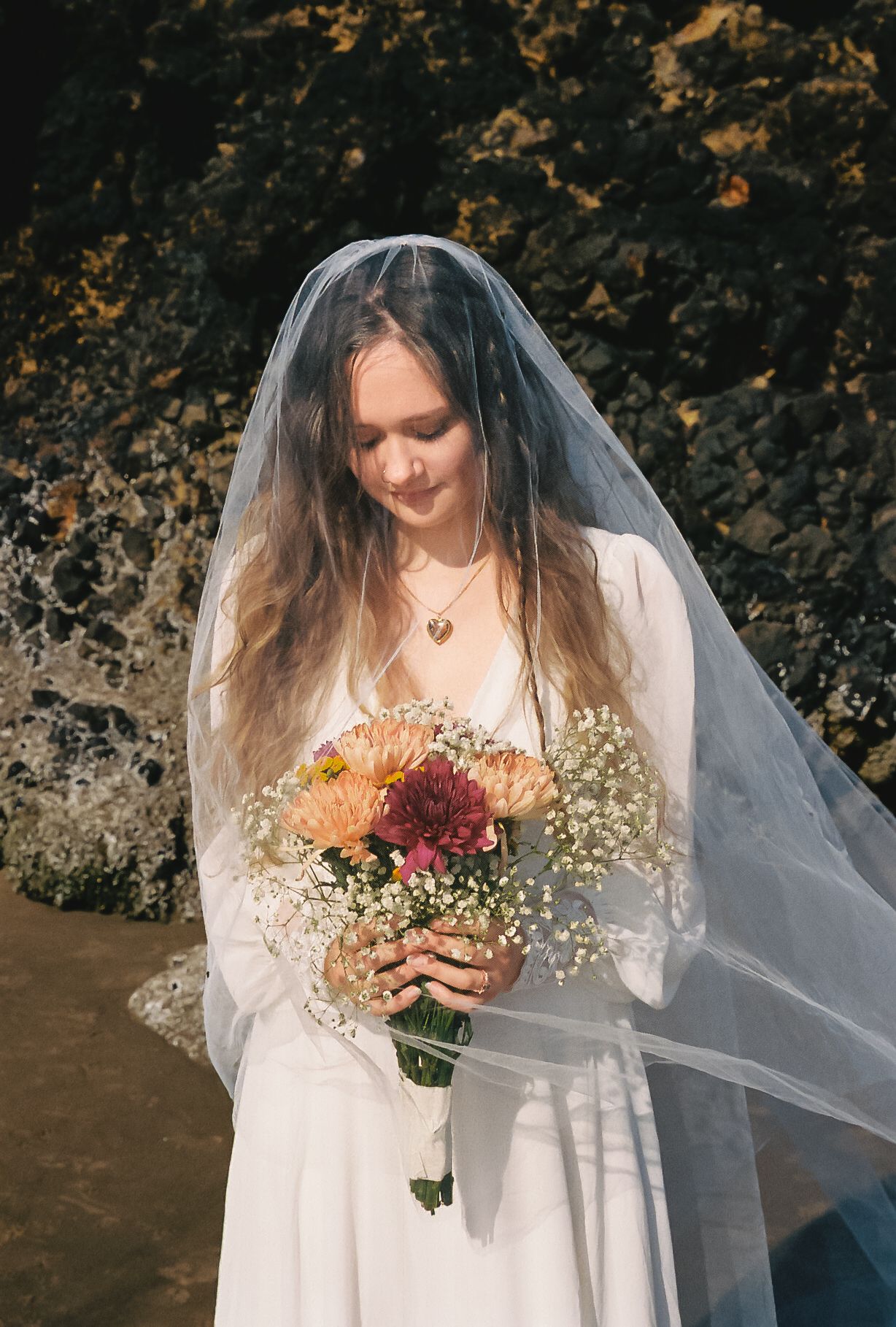 A bride in a white dress and veil looks down at her autumn-colored bouquet against a dark rocky backdrop.