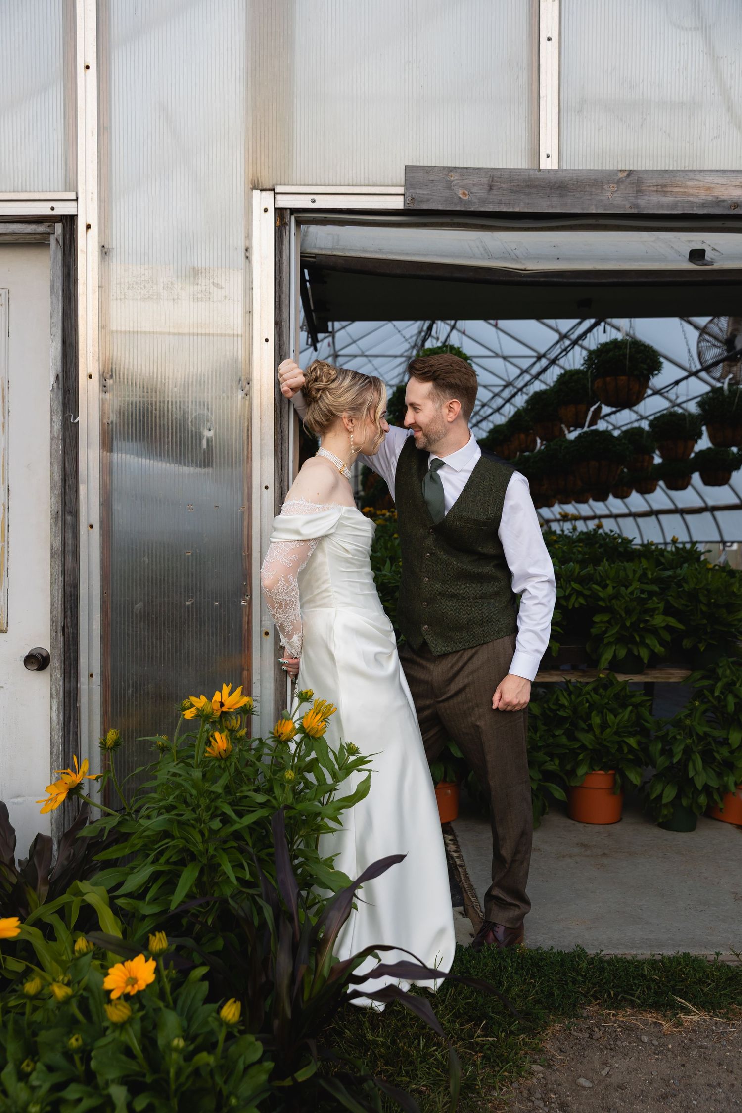 A couple in wedding attire share a romantic moment by a greenhouse doorway surrounded by yellow flowers.