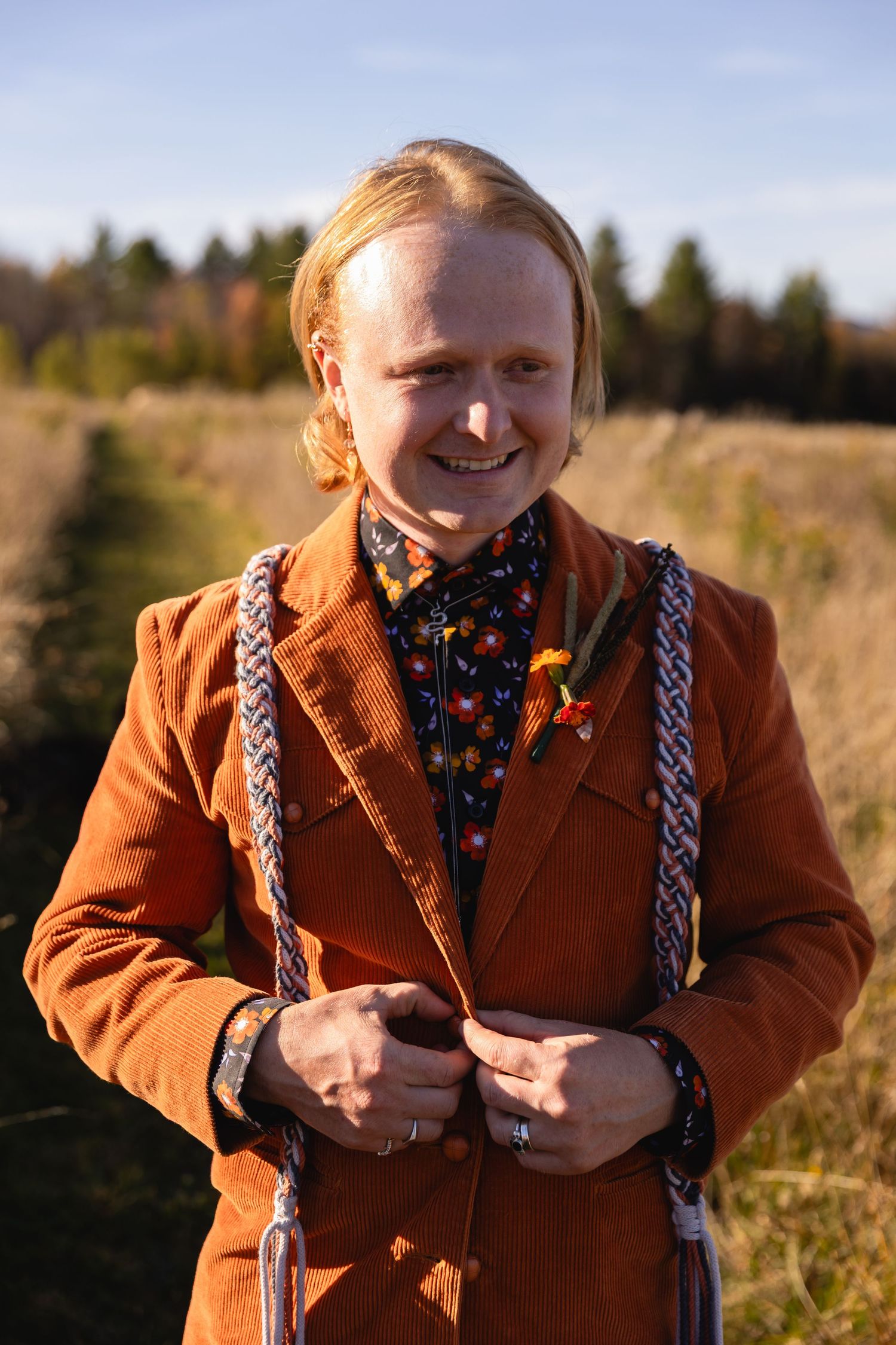 A person in a brown coat with braided hair smiles and poses in a sunny autumn field.