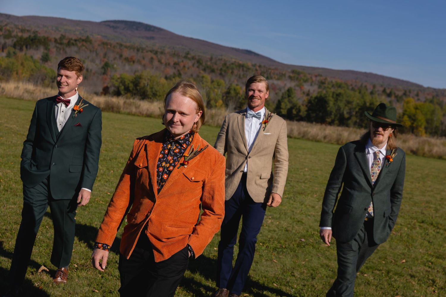 Wedding party walking in a line across a grassy field with mountains in the background on a sunny autumn day.
