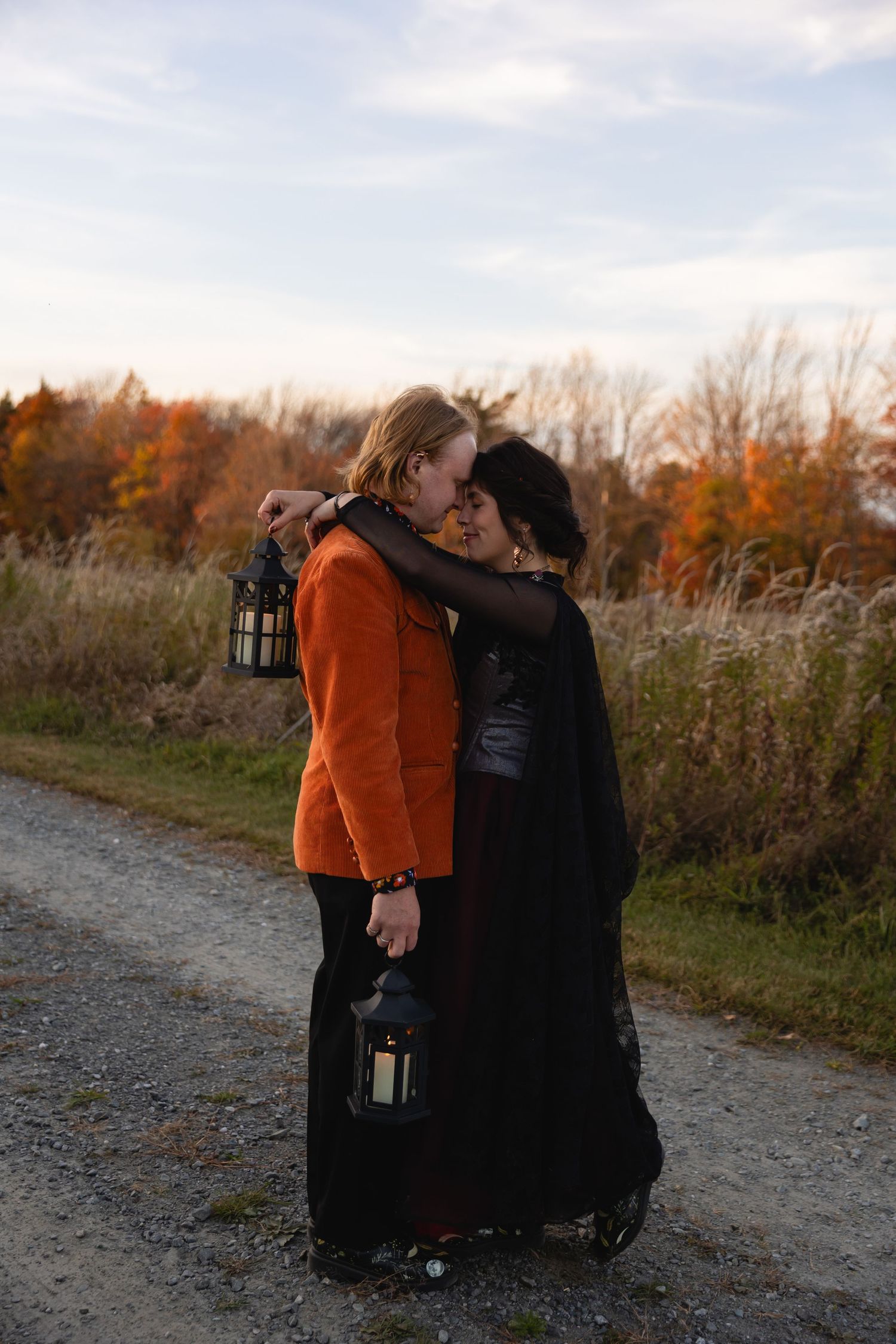 Two people share a romantic moment on a dirt path surrounded by autumn foliage while holding lanterns at dusk.