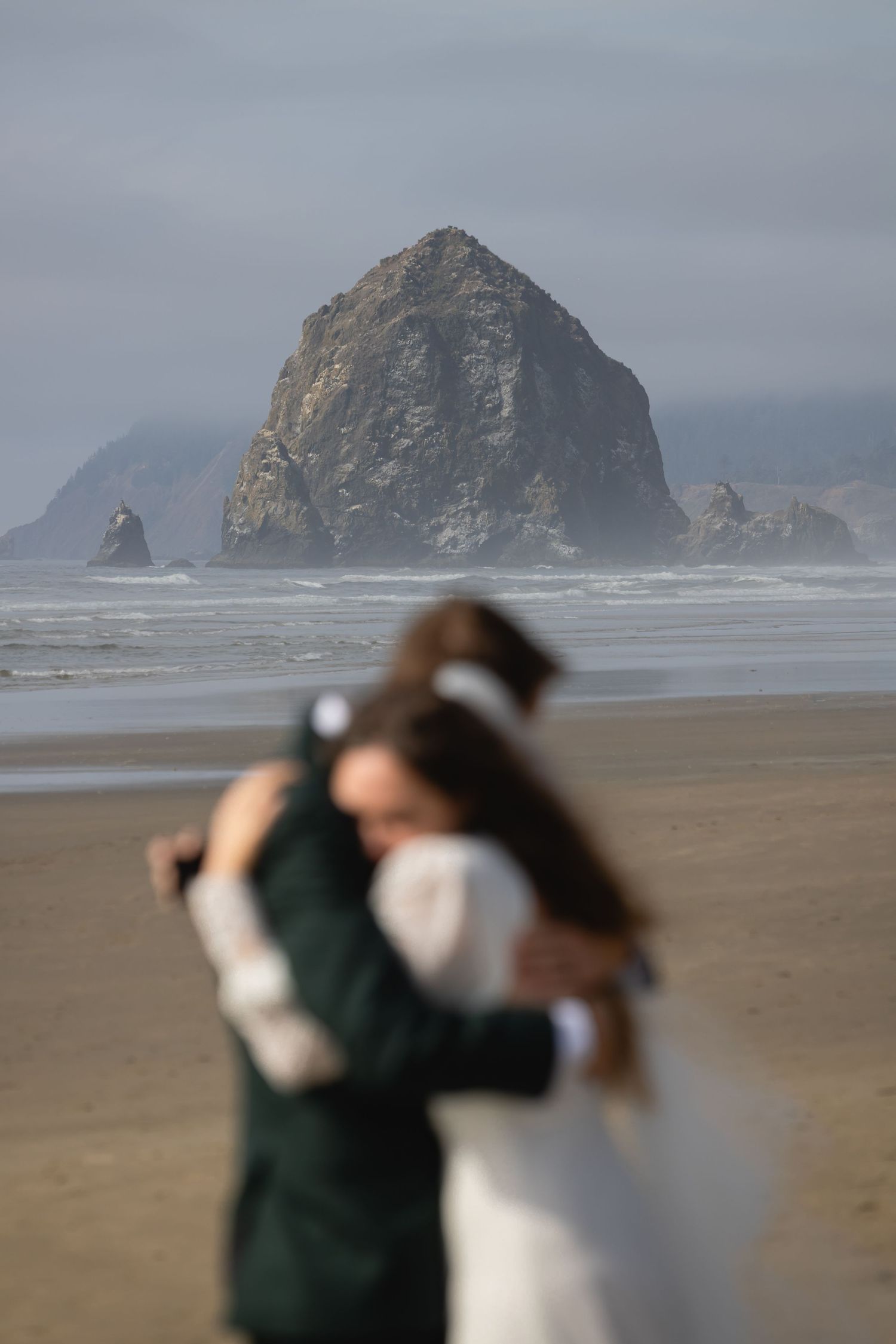 A couple embraces on Cannon Beach, Oregon with the iconic Haystack Rock formation looming in the misty background.