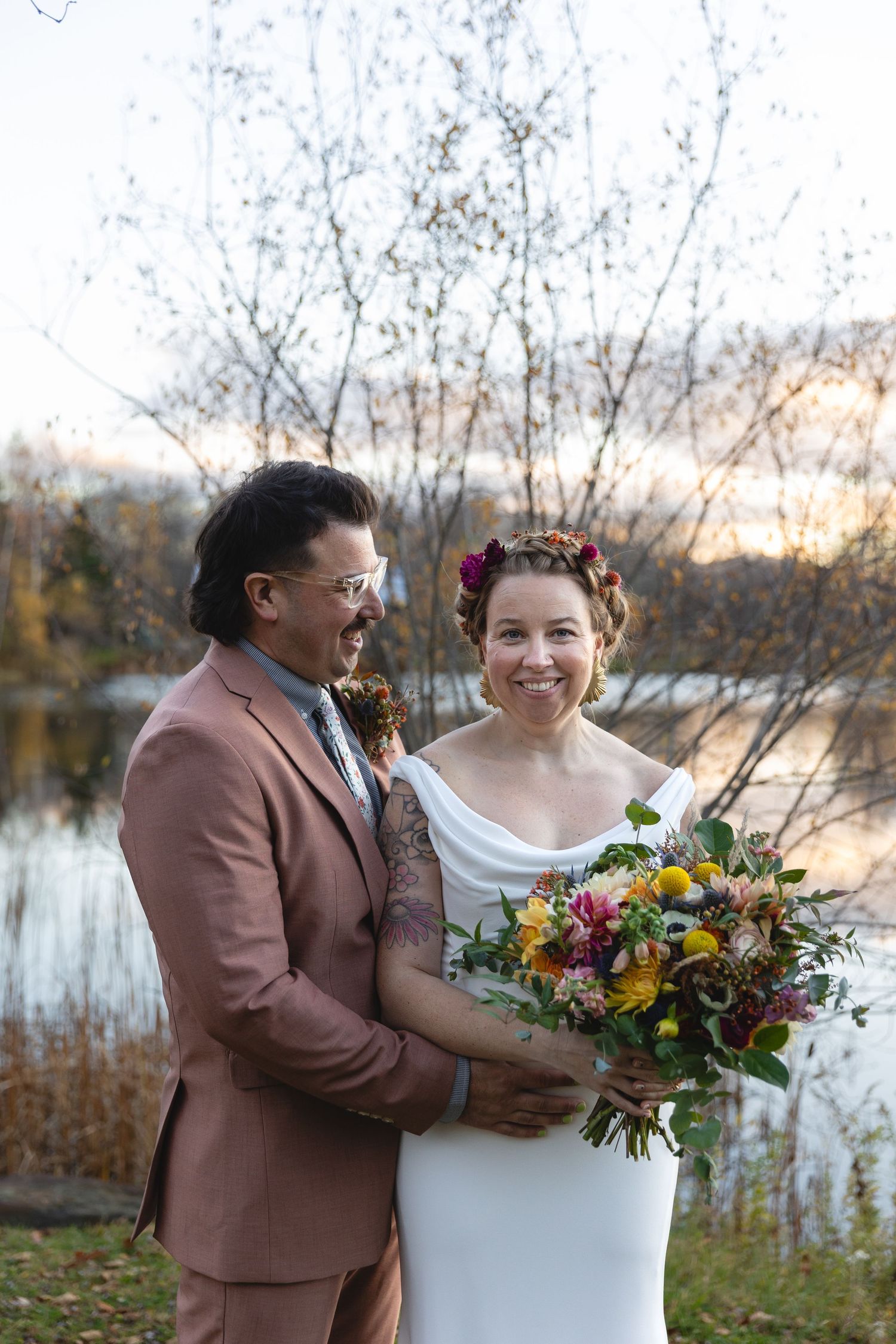 Couple poses with vibrant autumn bouquet during outdoor lakeside wedding.