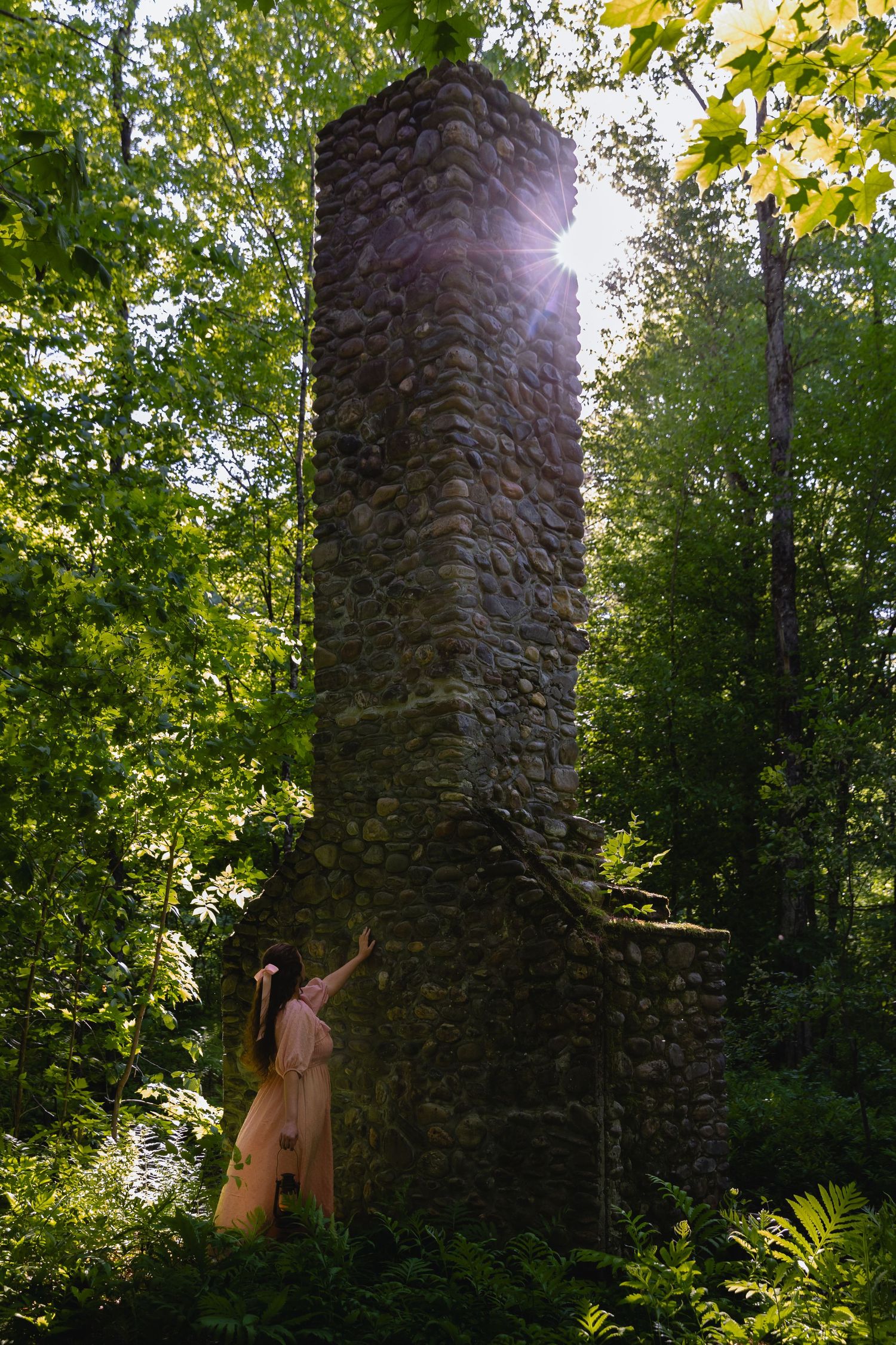 Woman in pink dress touching tall stone chimney tower with sunburst in green forest.