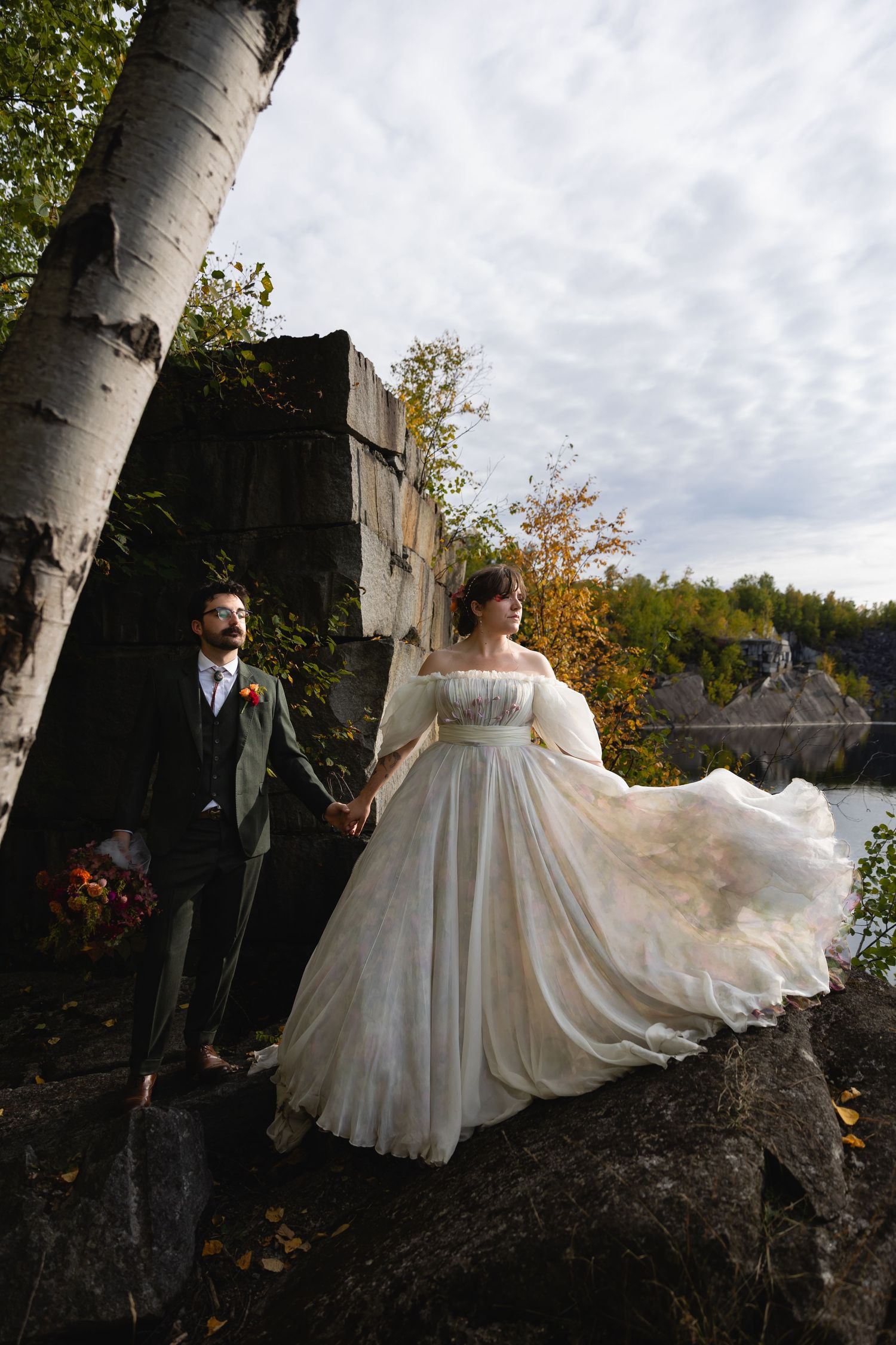 Bride in flowing white gown holds groom's hand near birch tree by quarry lake.