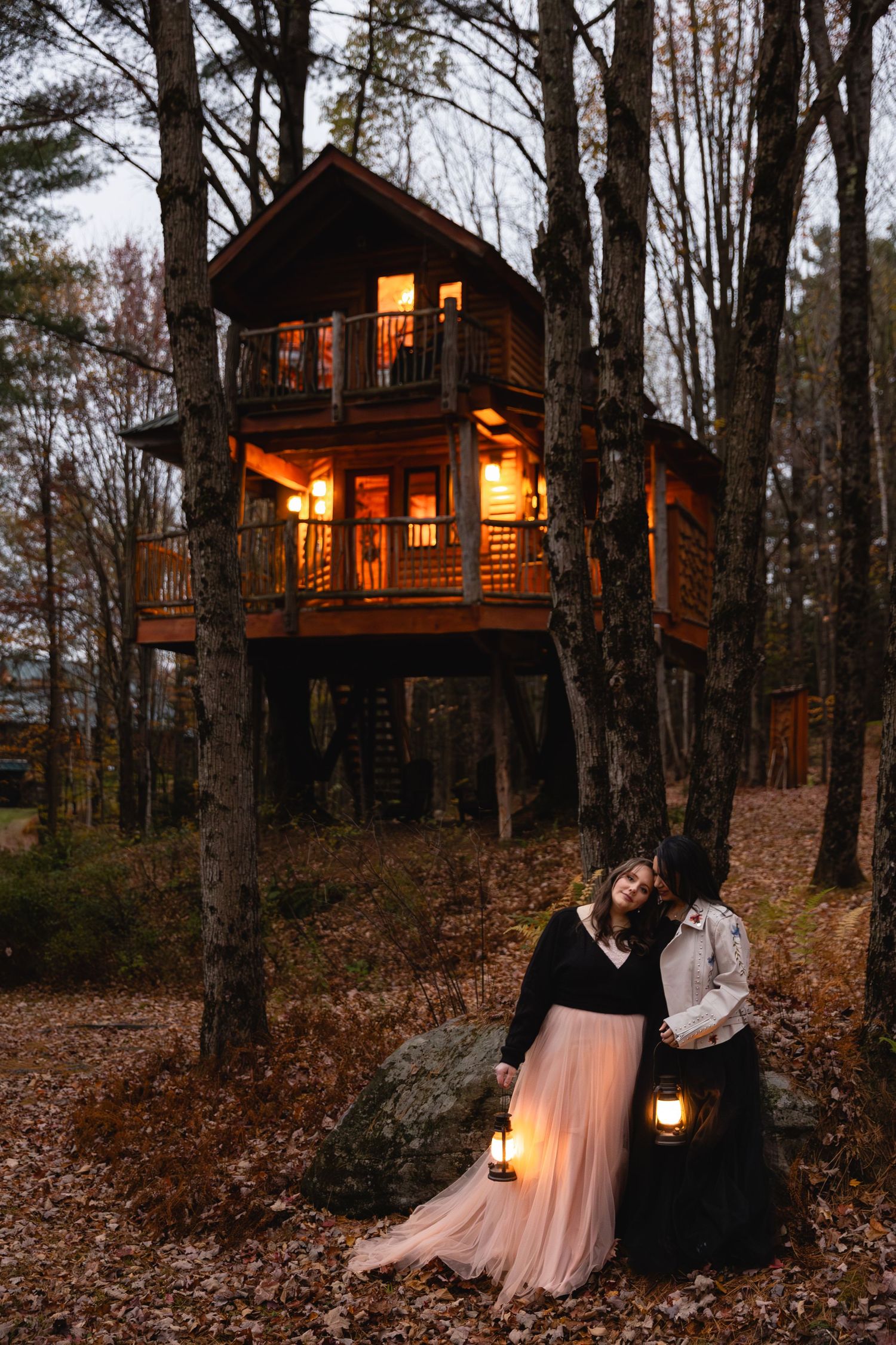 A couple embraces by lantern light in front of a glowing treehouse surrounded by autumn woods.