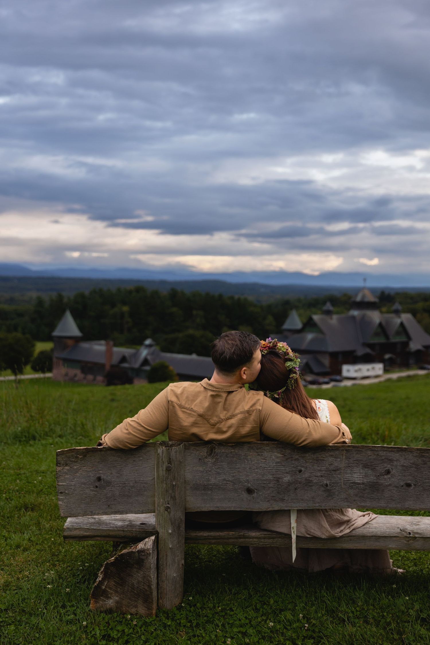 Couple sitting on wooden bench overlooking scenic countryside village under dramatic cloudy sky.